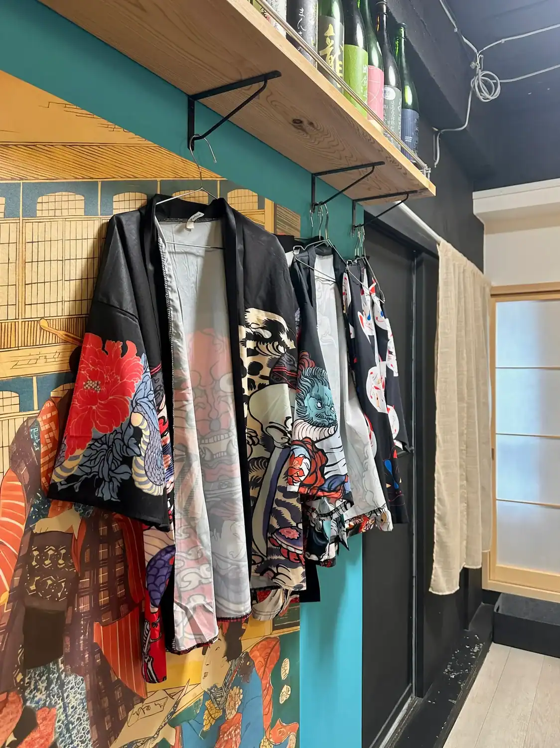Several colorful, patterned happi coat with bold Japanese designs hang on hooks beneath a wooden shelf holding sake bottles for a sushi making class in Tokyo