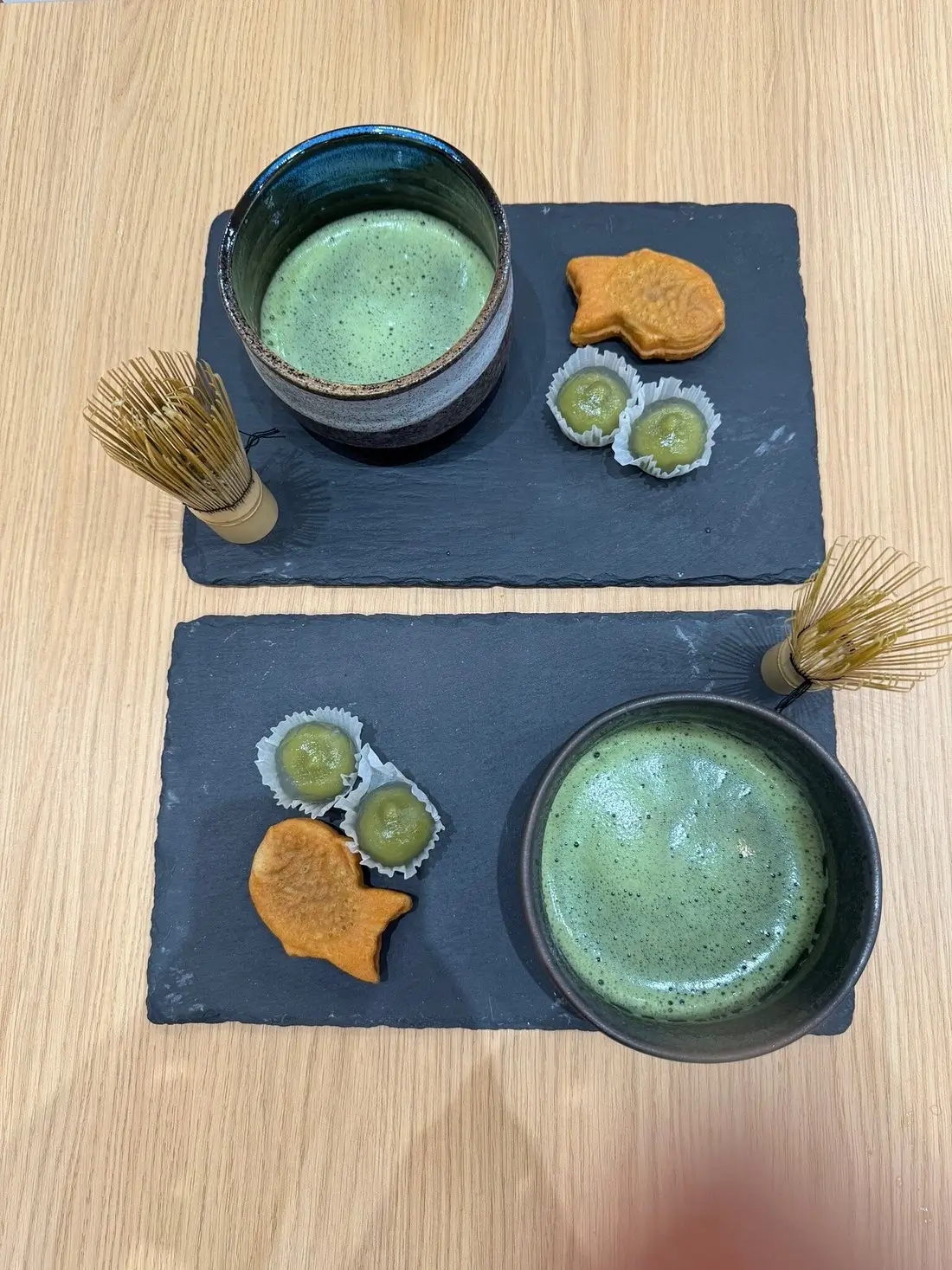 Two servings of matcha tea in ceramic bowls placed on black slate trays, each with small traditional Japanese sweets including fish-shaped cakes and mochi, arranged neatly on a wooden table during a matcha-making class.