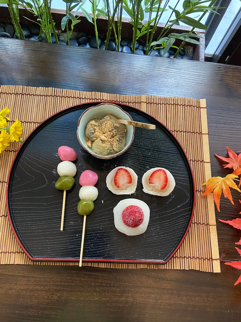 Traditional Japanese sweets including ichigo daifuku (strawberry mochi), dango skewers in pink, white, and green, and warabi mochi topped with kinako powder, served on a black lacquer tray with autumn decor during a mochi-making class.