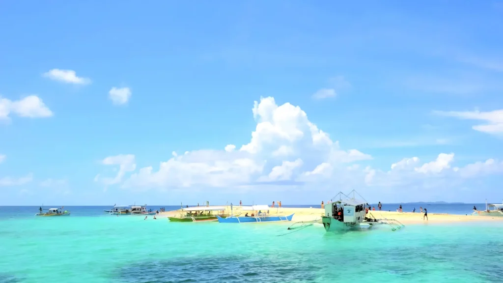 A small sandbar surrounded by clear turquoise water in Siargao, with several boats docked and people walking along the shoreline under a bright blue sky.