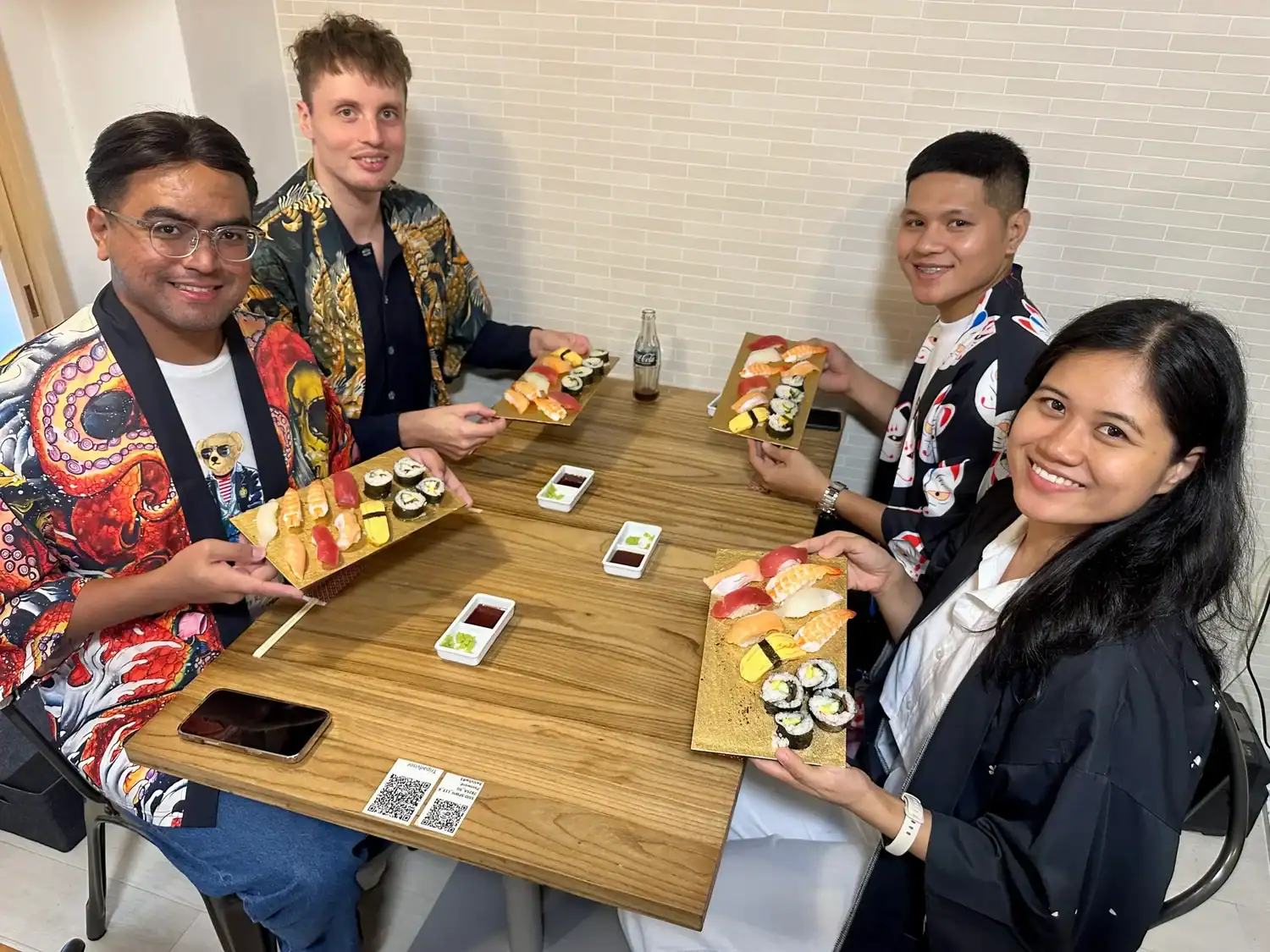 Four people smile while seated at a wooden table, each holding a tray of assorted sushi including nigiri and maki rolls during a sushi class in Tokyo