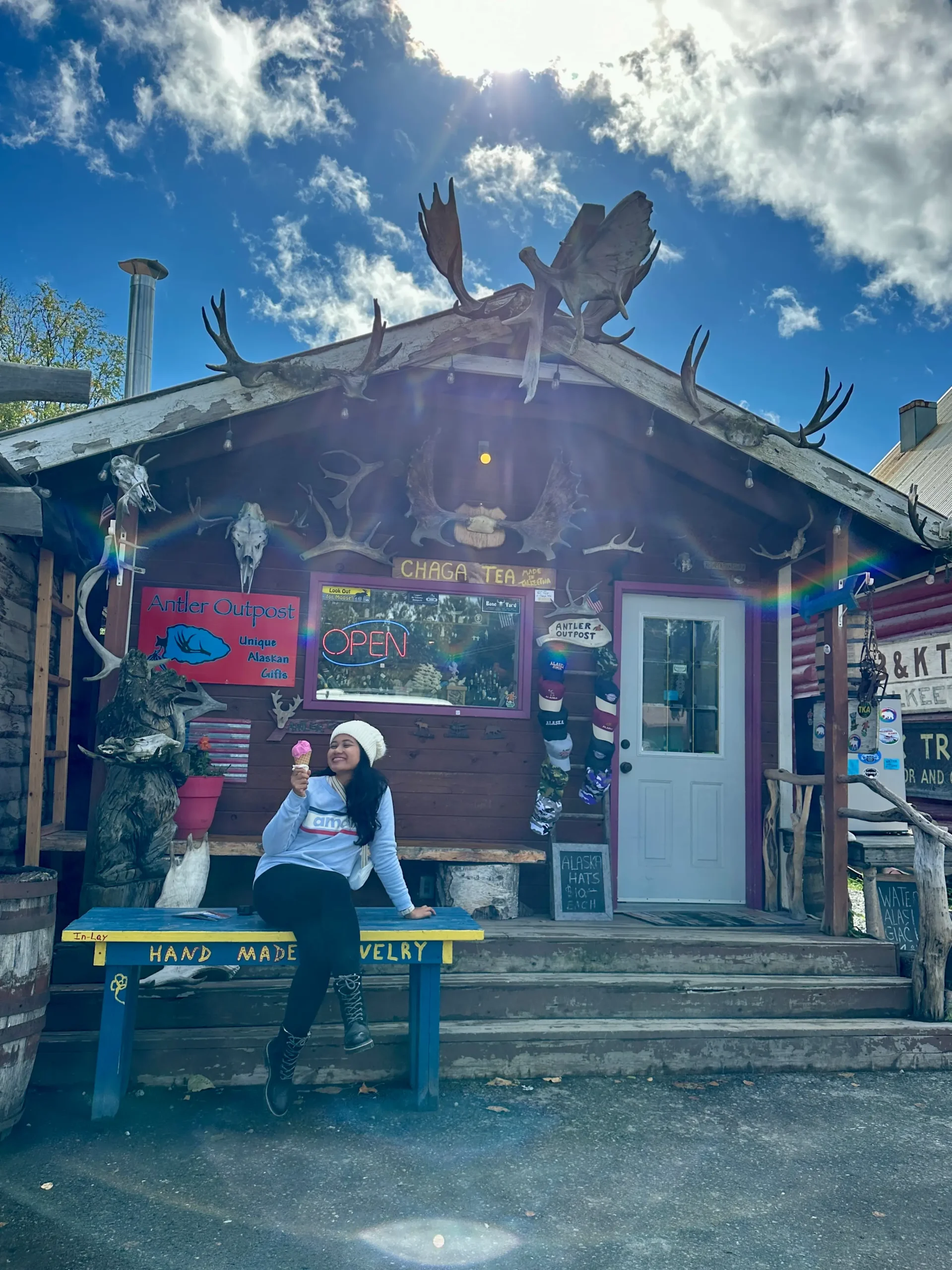 A woman sitting on a bench and enjoying a pink fireweed ice cream cone in front of a red cabin-style shop decorated with antlers and a “CHAGA TEA” sign as one of the best things to do in Talkeetna Alaska