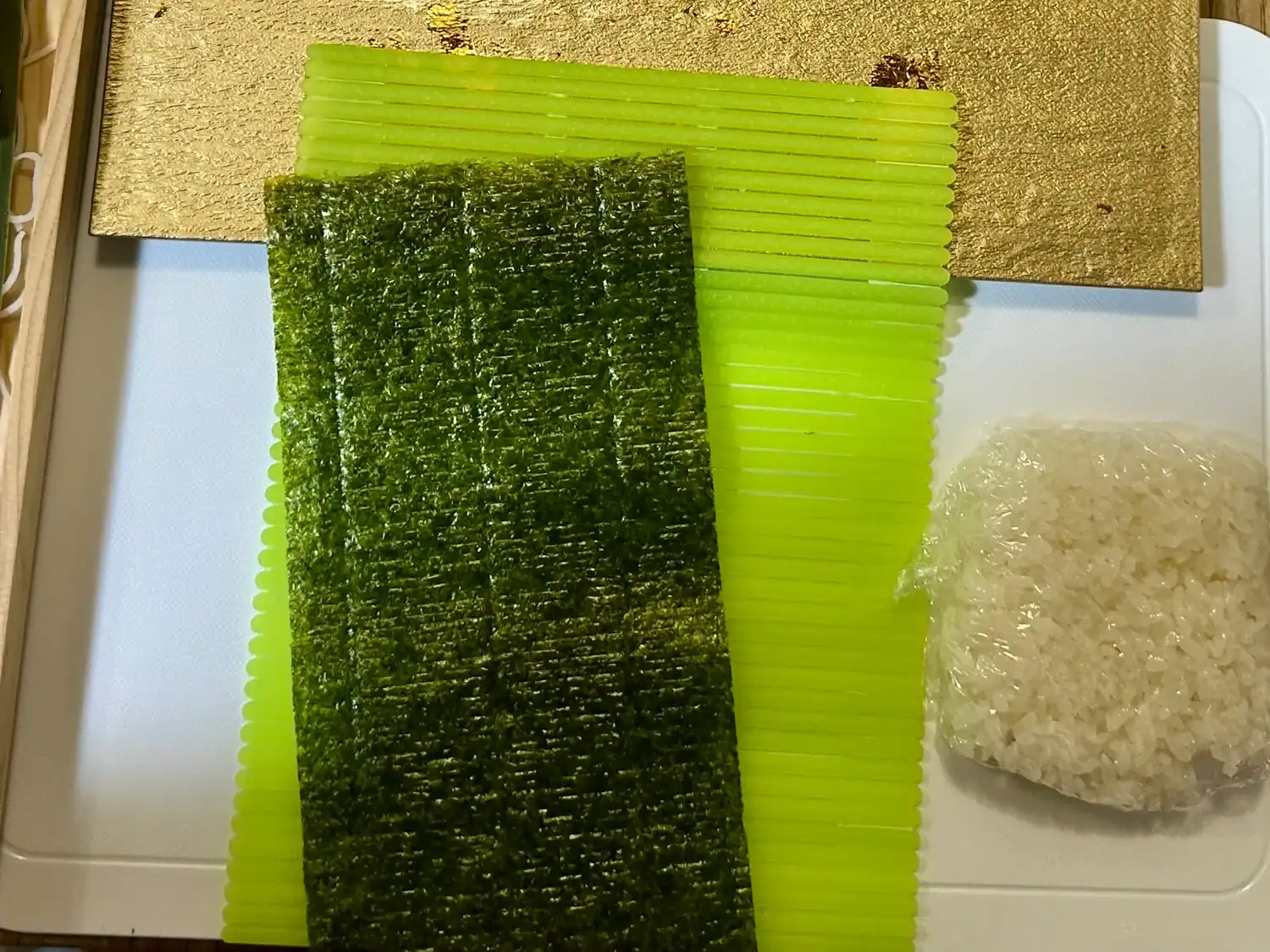 A sushi-making setup featuring a sheet of nori on a plastic green bamboo mat, a portion of rice wrapped in plastic wrap, and a gold plate partially visible in the background.