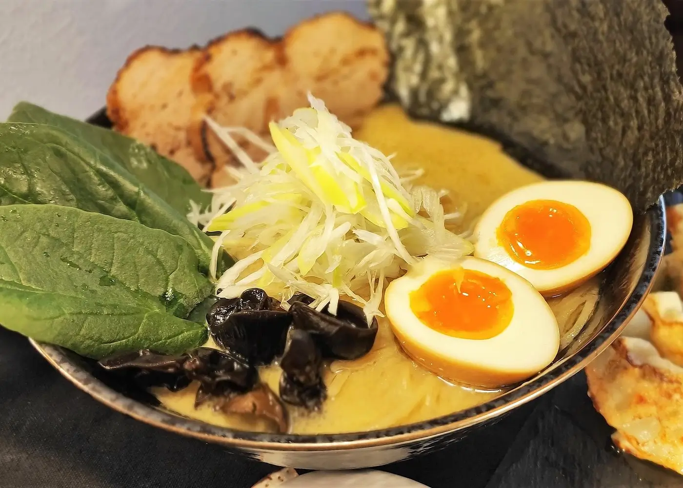 A bowl of Japanese ramen topped with soft-boiled eggs, sliced chashu pork, spinach leaves, bamboo shoots, mushrooms, shredded green onions, and a sheet of seaweed, served with a side of gyoza dumplings.