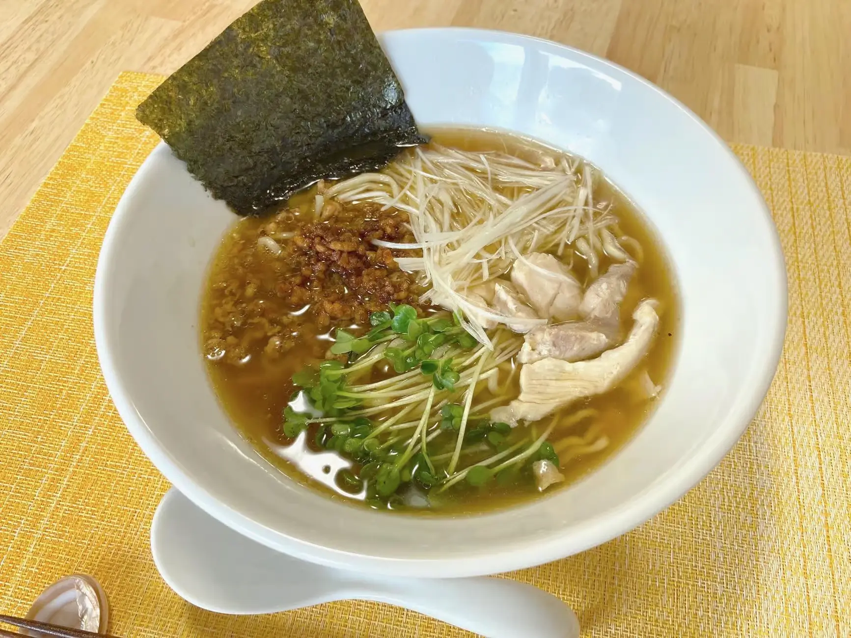 A bowl of ramen with chicken slices, sprouts, fried garlic, and a sheet of nori, served in a white bowl on a yellow placemat.