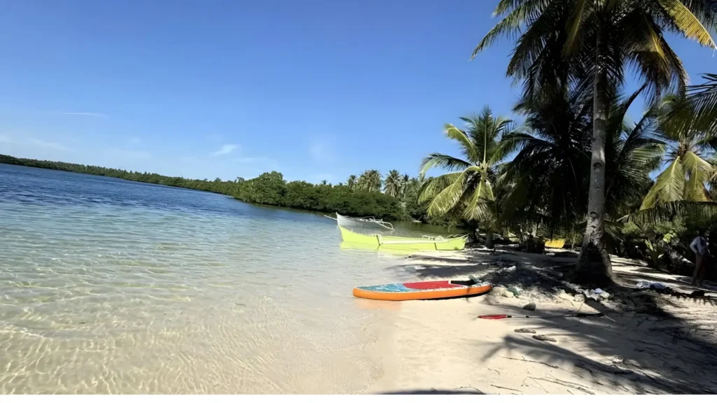 A quiet shoreline at Secret Beach in Siargao with clear shallow water, a paddleboard, and small boats shaded by palm trees.