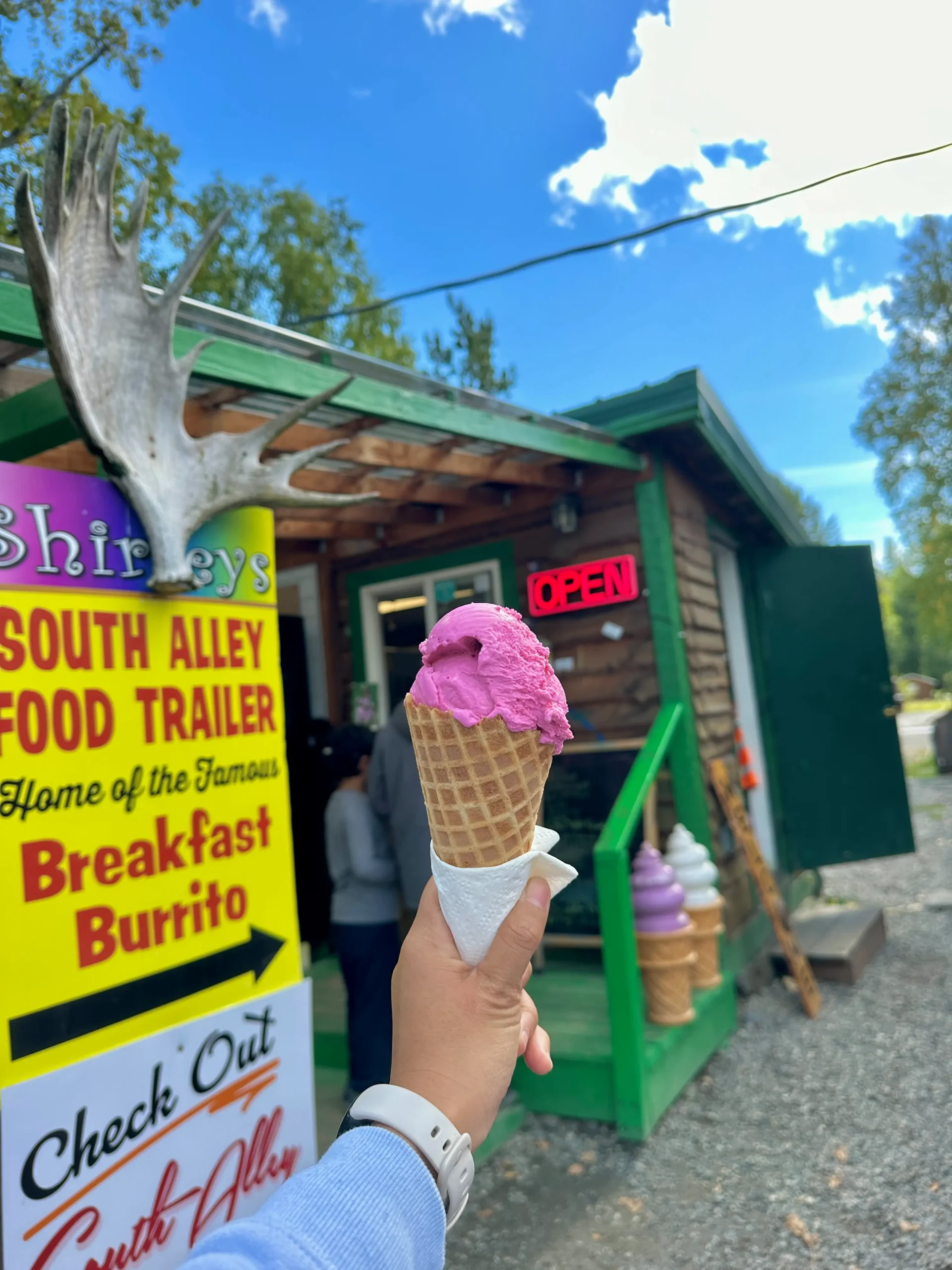 Hand holding a pink fireweed ice cream cone in front of Shirley’s Ice Cream, a small wooden shop with green accents and large ice cream cone sculptures by the entrance.