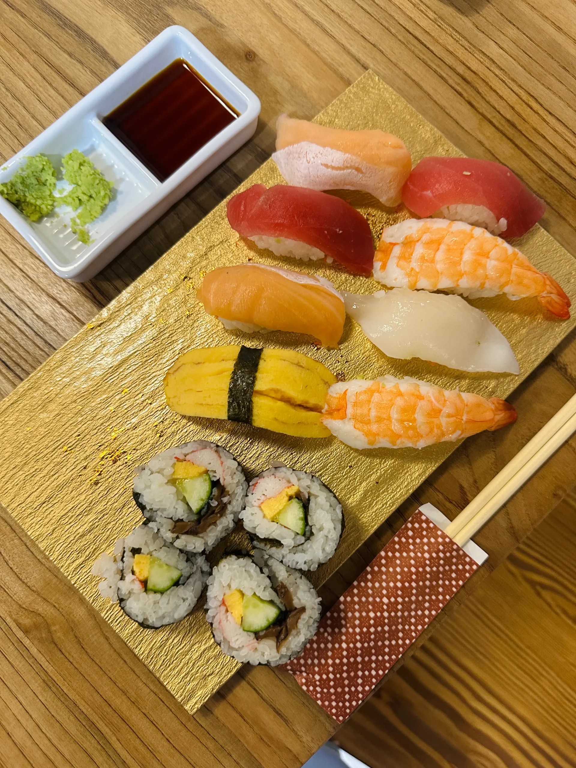 A close-up of a golden tray with assorted sushi including shrimp, tuna, tamago, and cucumber maki rolls, placed beside a rectangular dish of soy sauce and wasabi and a pair of chopsticks in a red wrapper during a sushi class in tokyo
