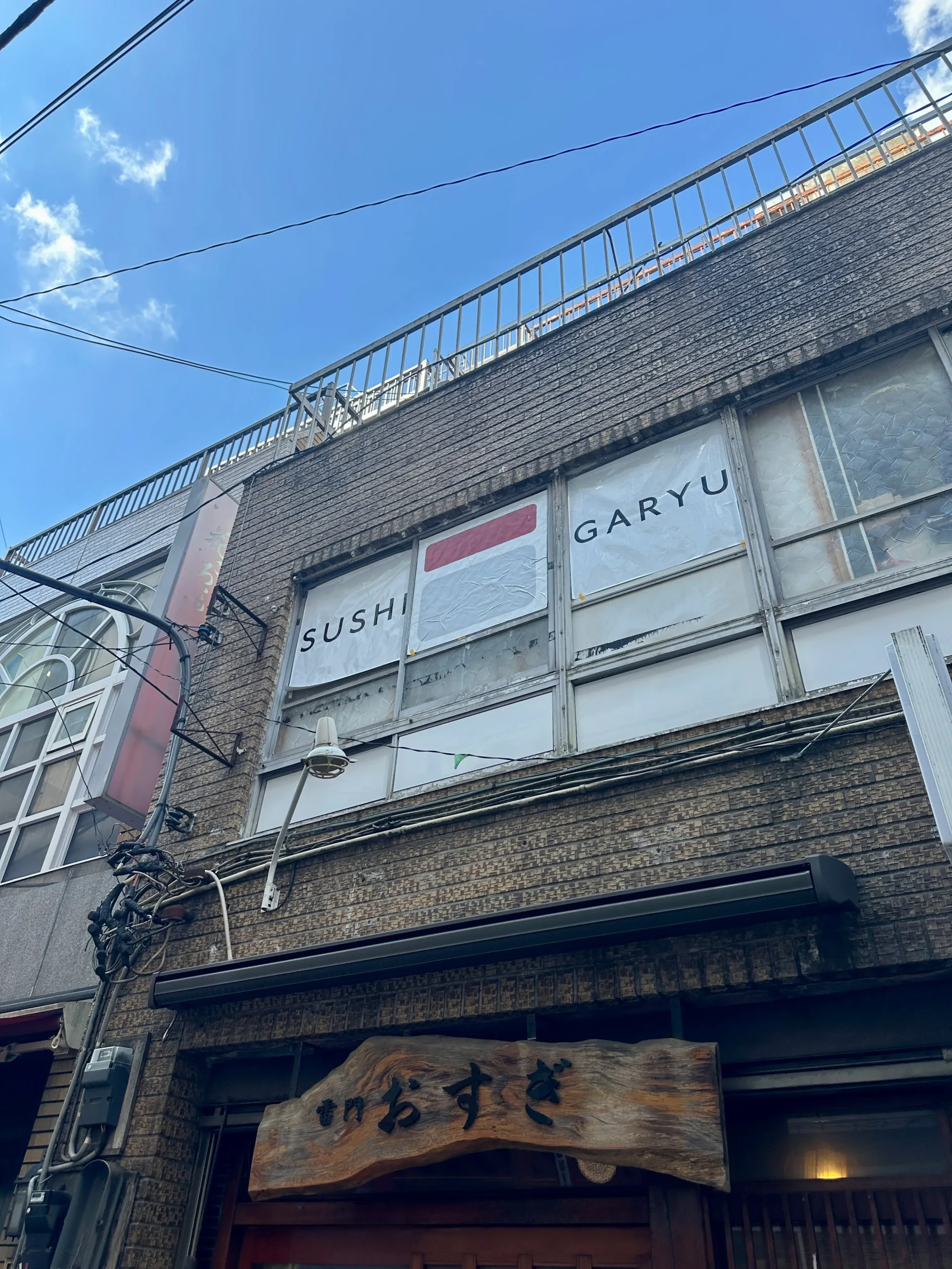 Exterior of a rustic building with a sign reading “SUSHI GARYU” in the window above a wooden sign with Japanese characters, set against a clear blue sky.