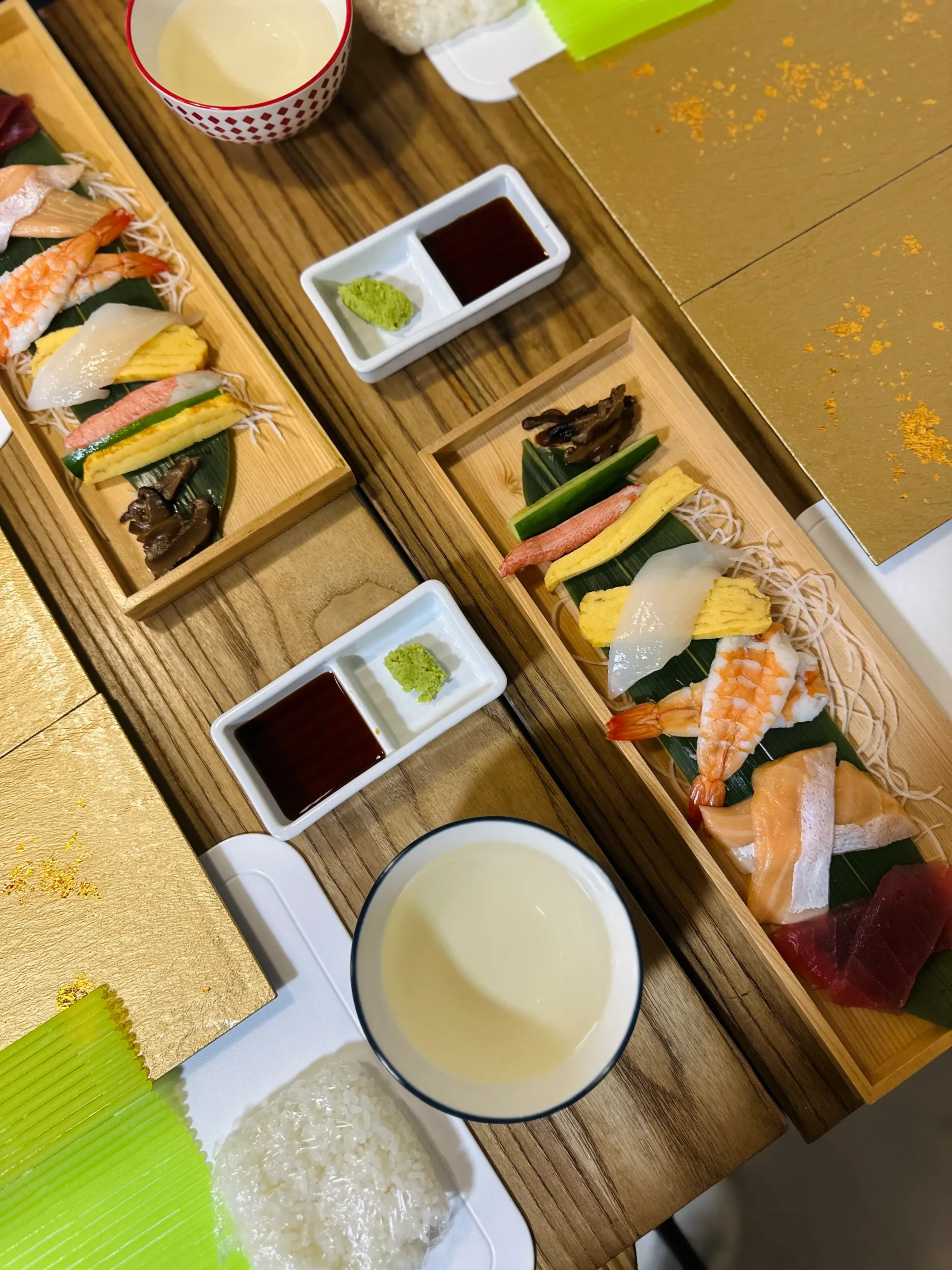 An overhead view of wooden trays filled with sushi ingredients like raw fish, tamago, vegetables, and imitation crab, alongside soy sauce, wasabi, and sake cups, arranged for a sushi making class in Japan