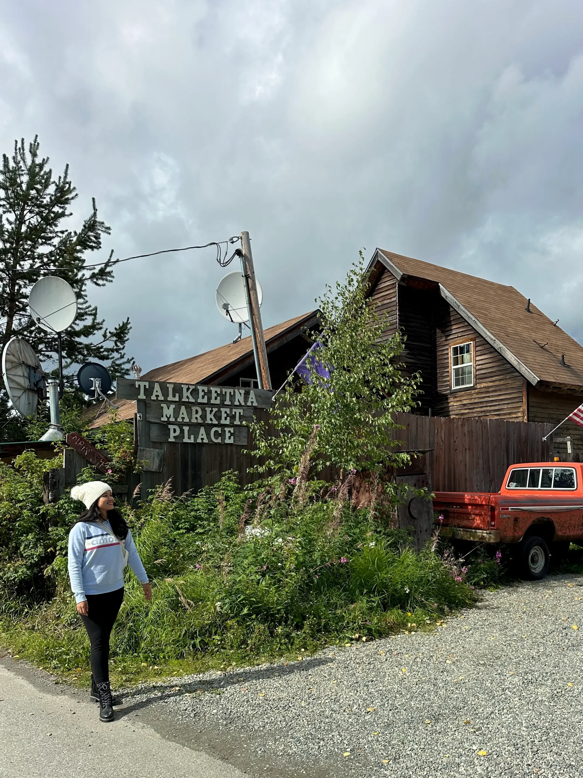 A woman standing on a sidewalk beside a cabin-style store with the sign, "Talkeetna Market Place," as well as a red pickup truck.