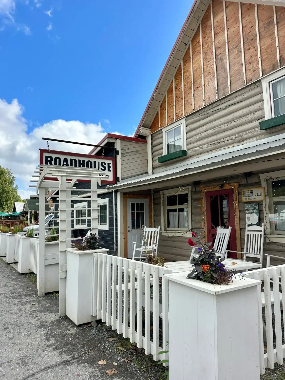 Front facade of the Talkeetna Roadhouse, one of the most historic places to stay in Talkeetna, featuring a white picket fence, rocking chairs, and a rustic log cabin exterior under a clear blue sky.