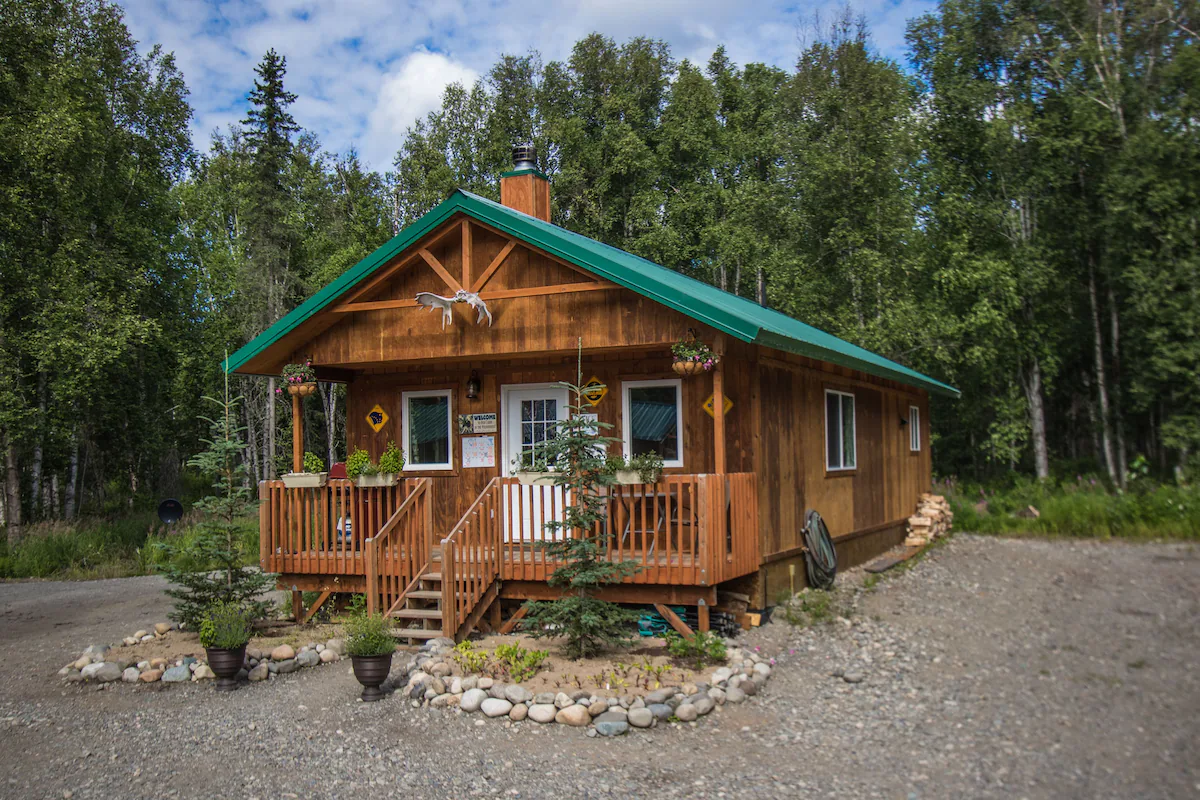 One of the cabins in Talkeetna Wilderness Lodge and Cabin Rentals, complete with its front deck, green-painted roof, and surrounding forest.