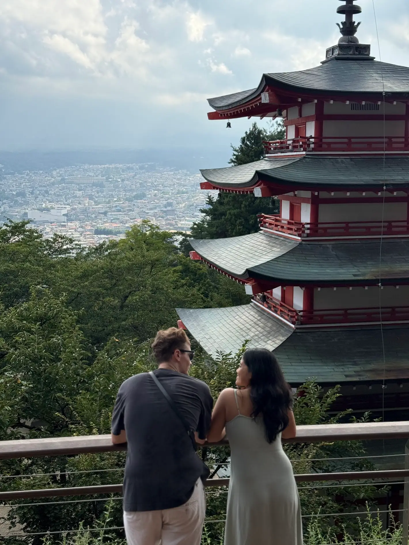 A couple standing at the lookout near Chureito Pagoda in Kawaguchiko, facing the city view and the pagoda structure on a cloudy day.