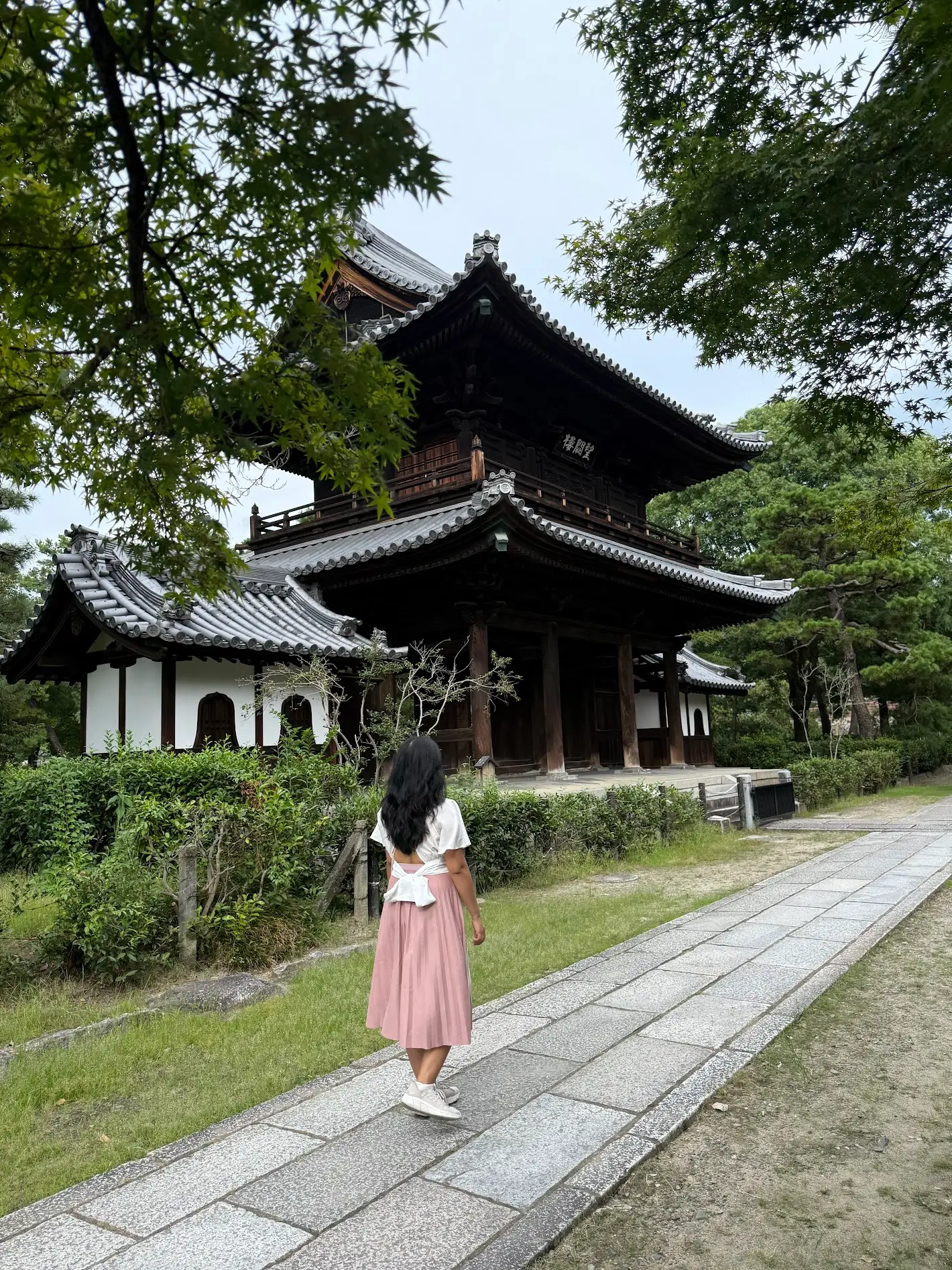 A woman walks on a stone path beside a traditional wooden temple surrounded by trees.