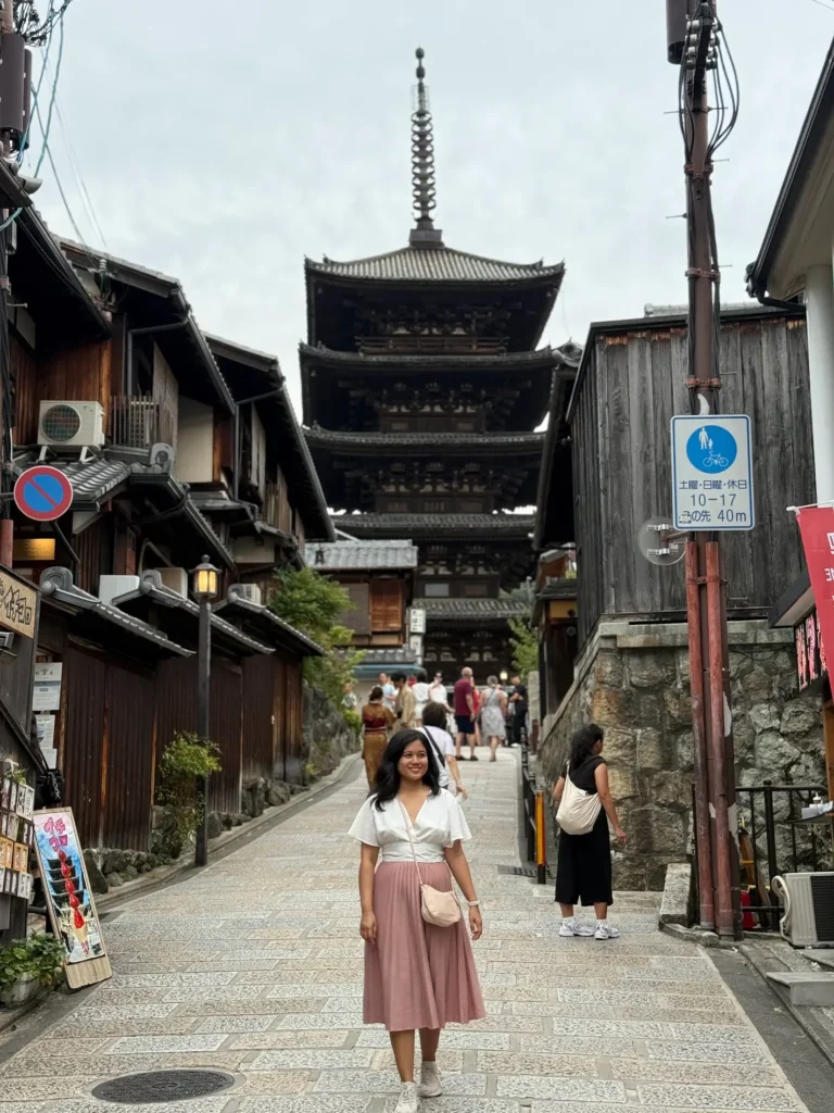 A woman walks along a stone street with wooden buildings and a five-story pagoda in the background.