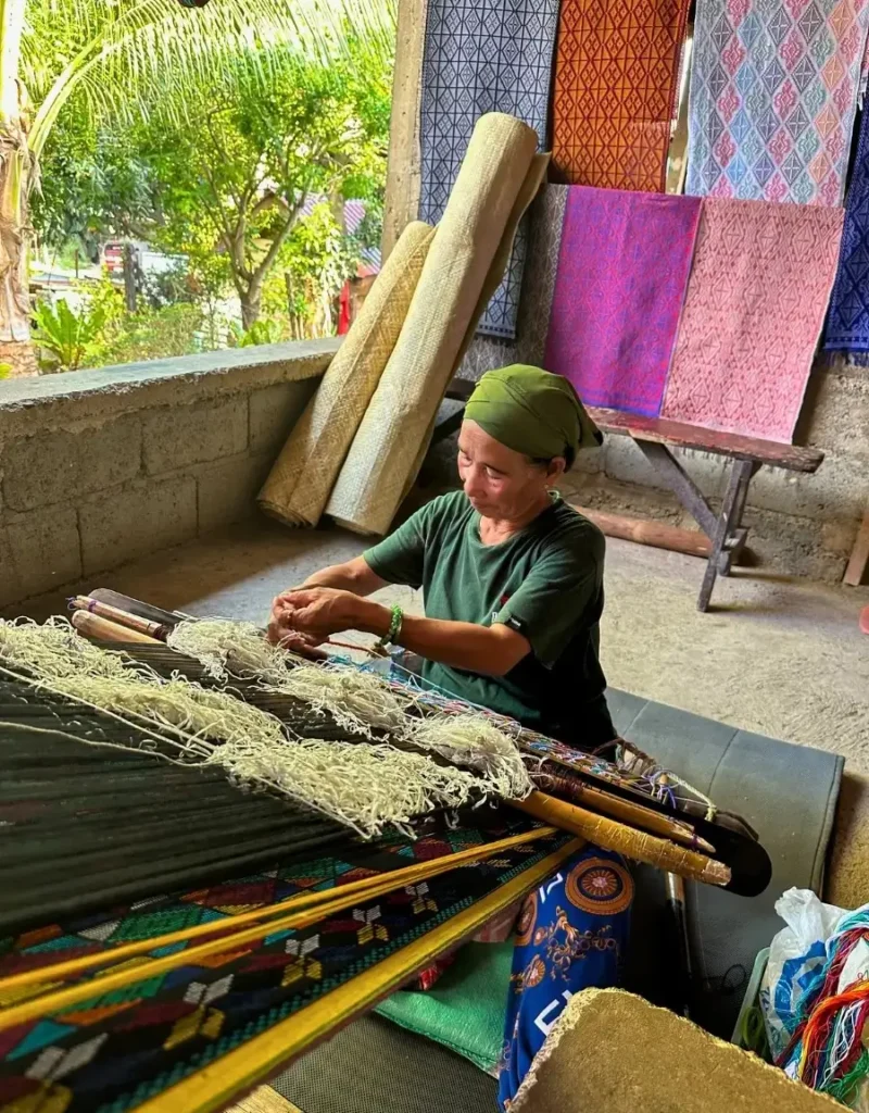 An elderly woman from the Yakan Village in Zamboanga sits in front of a traditional weaving equipment, manually piecing together strands of fabric to form colorful cloths.