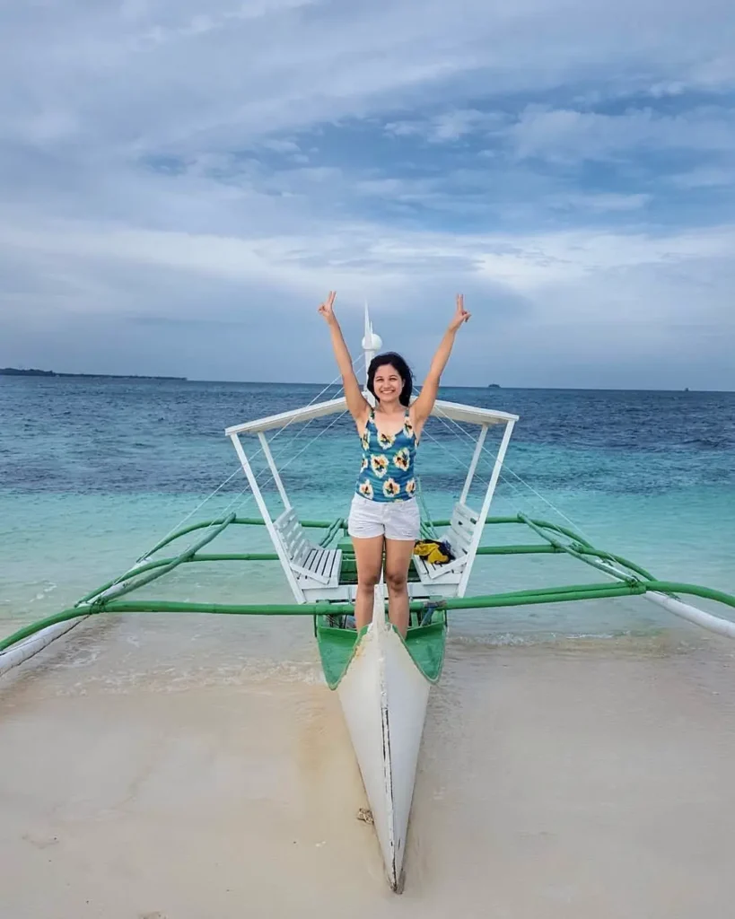 A woman standing at the front of a small boat on a sandy shore, smiling and raising both arms, with clear blue water in the background.