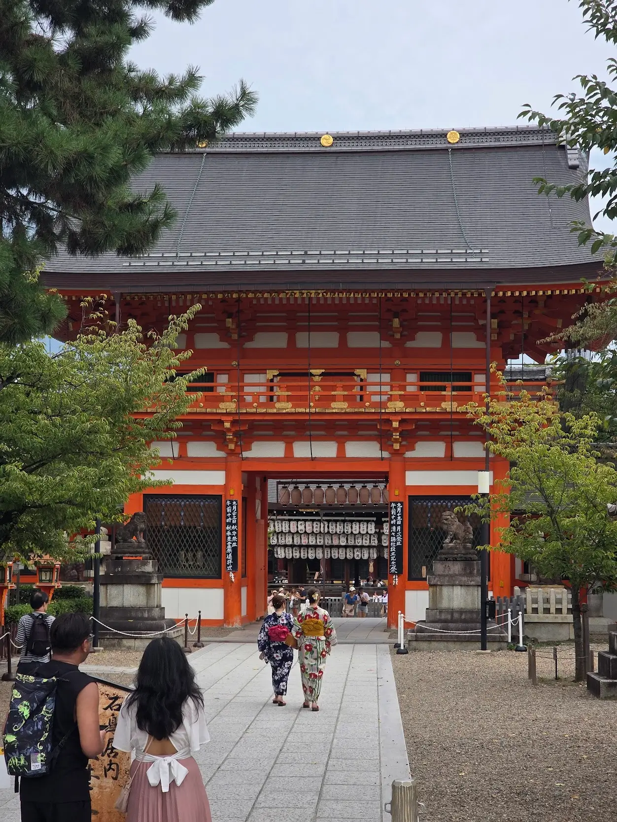 A bright orange shrine gate in Kyoto with visitors walking inside, including two people wearing yukata.