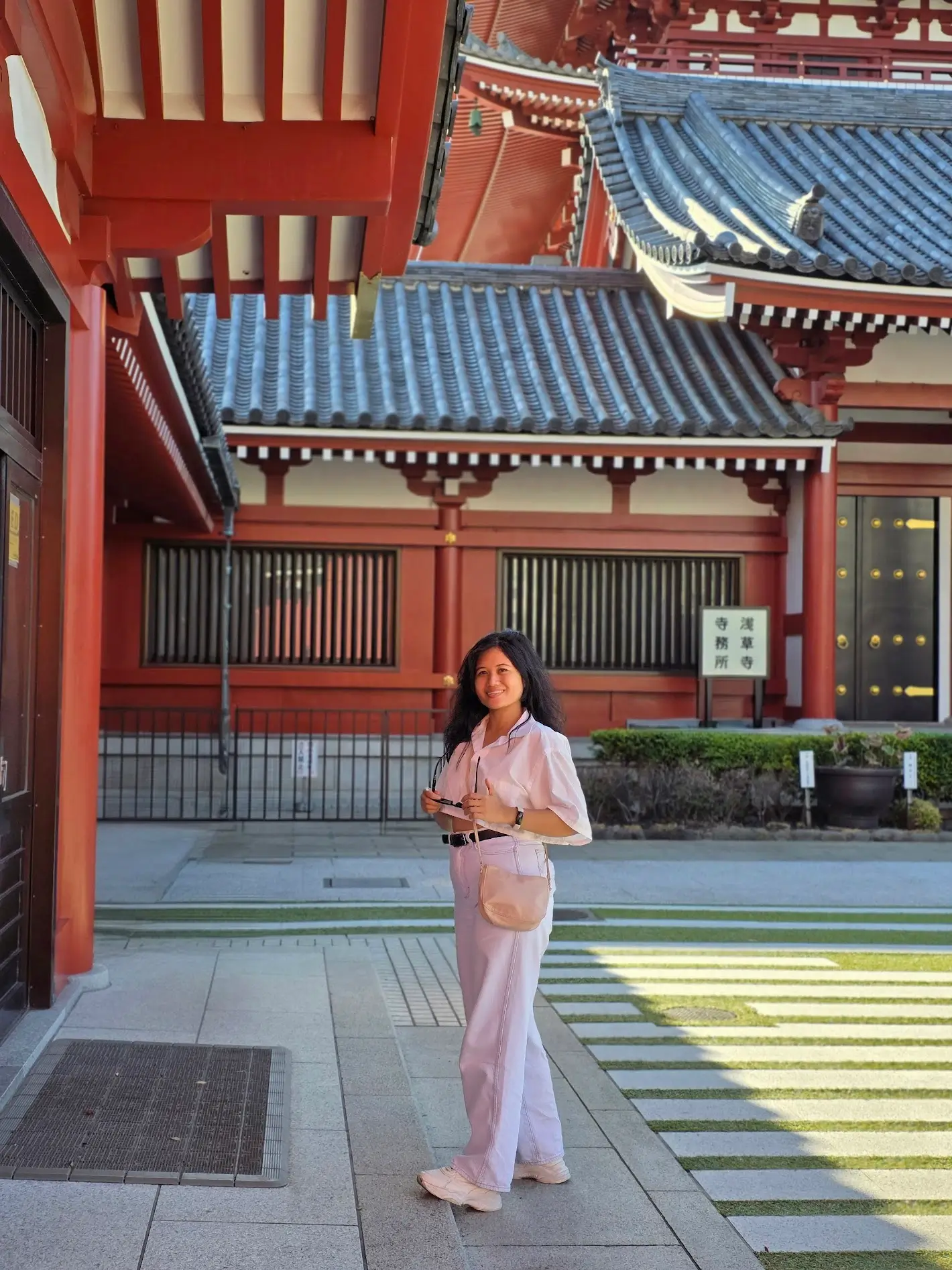 A woman stands in a temple courtyard wearing a light outfit, smiling toward the camera, with red wooden beams and tiled roofs behind her.