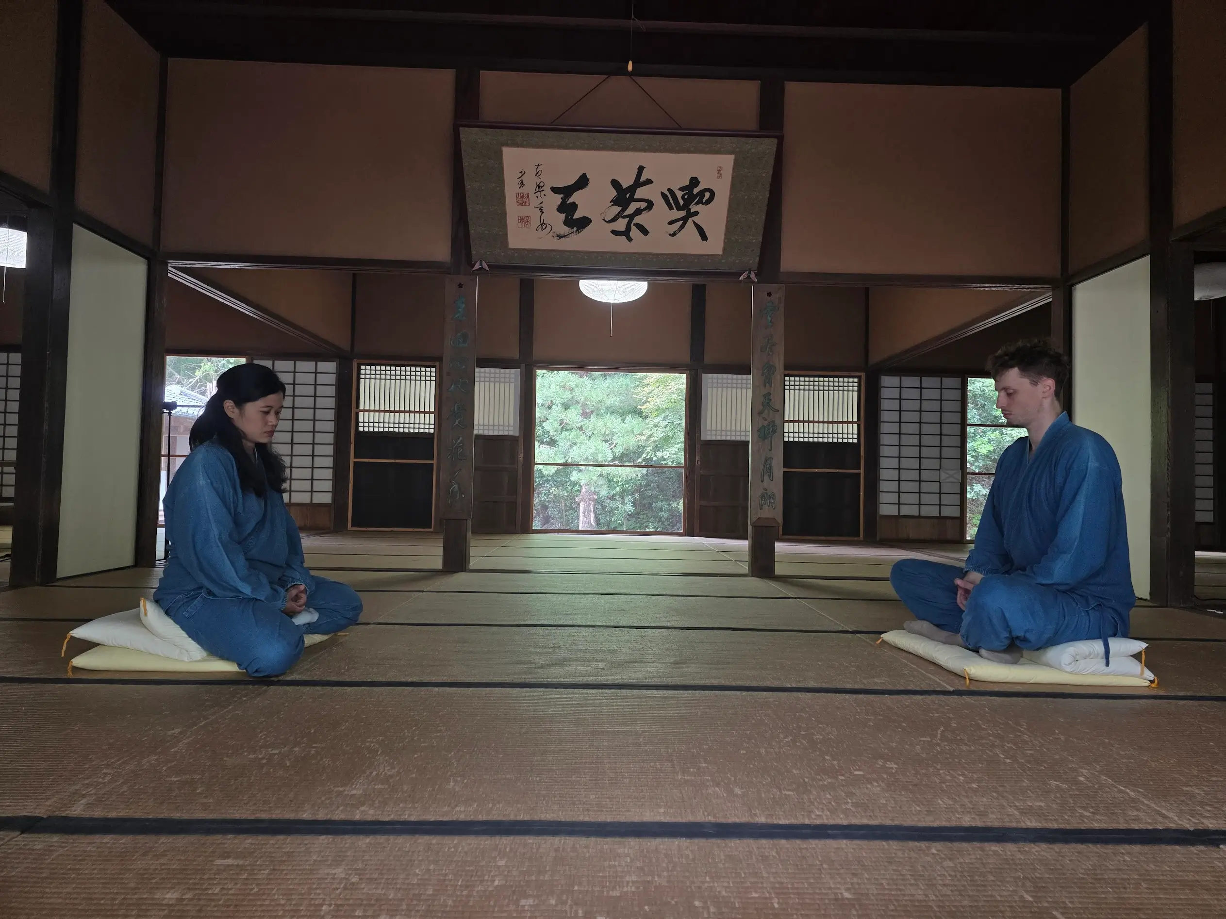 Two people in blue temple robes sit facing each other on cushions in a tatami room at Manpuku-ji Temple, practicing silent zazen meditation. Doing non-touristy things to do in Kyoto.