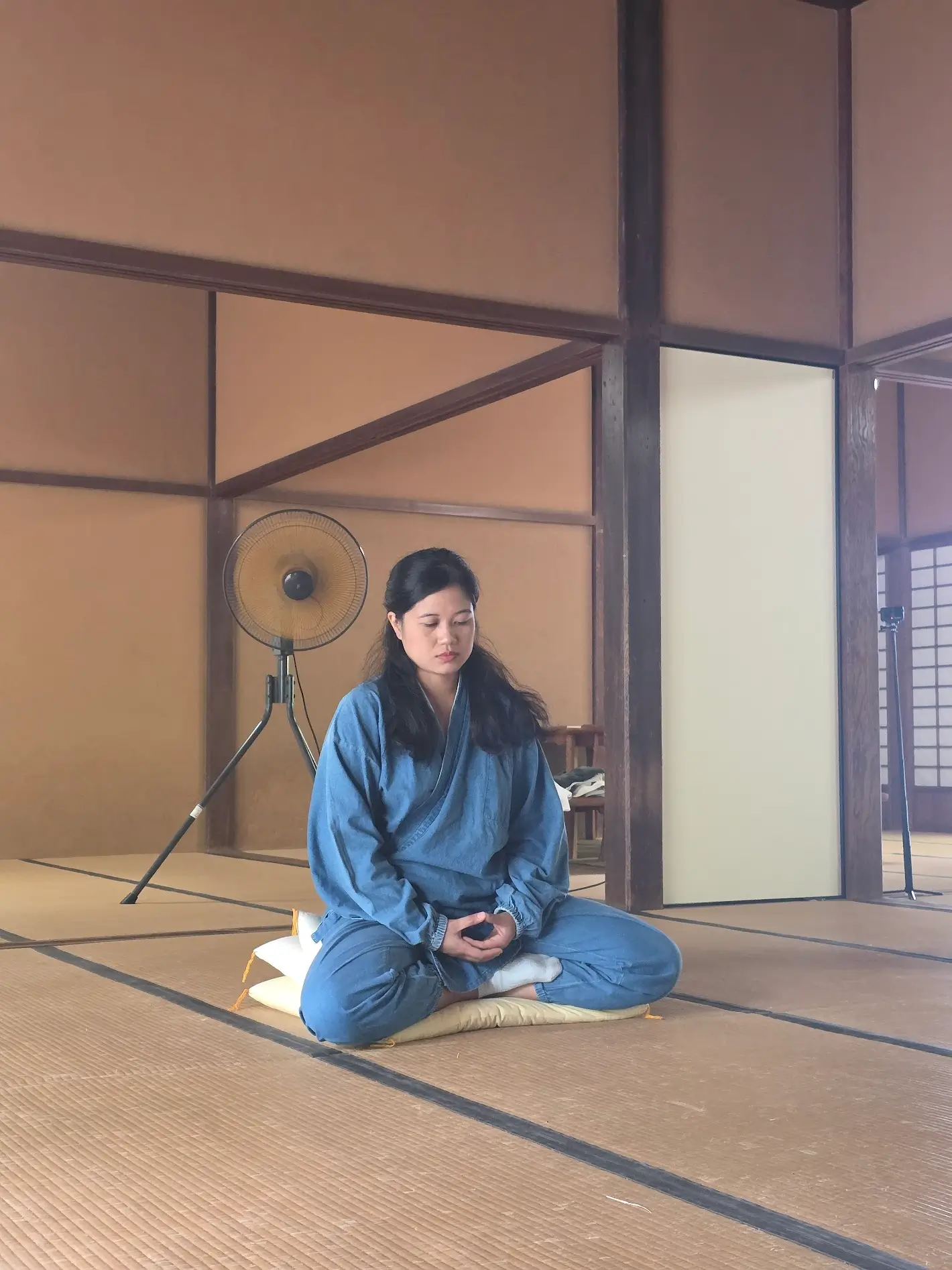 A woman in blue temple robes sits cross-legged on a cushion inside a tatami room at Manpuku-ji Temple, practicing quiet zazen meditation.