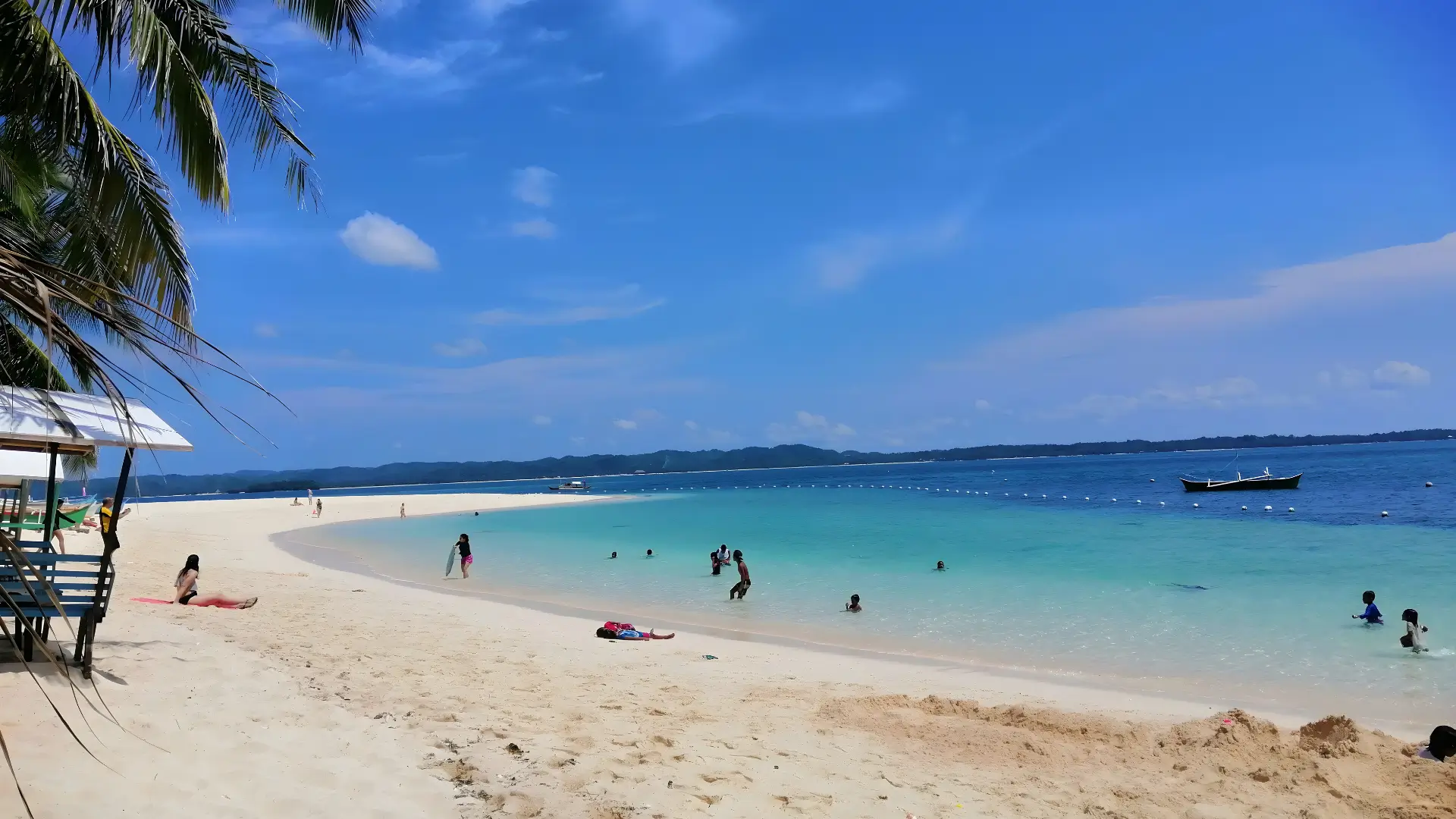 A quiet white sand beach with clear blue water at Alegria Beach in Siargao. A few people are swimming and relaxing along the shoreline under sunny weather. Best beaches in Siargao