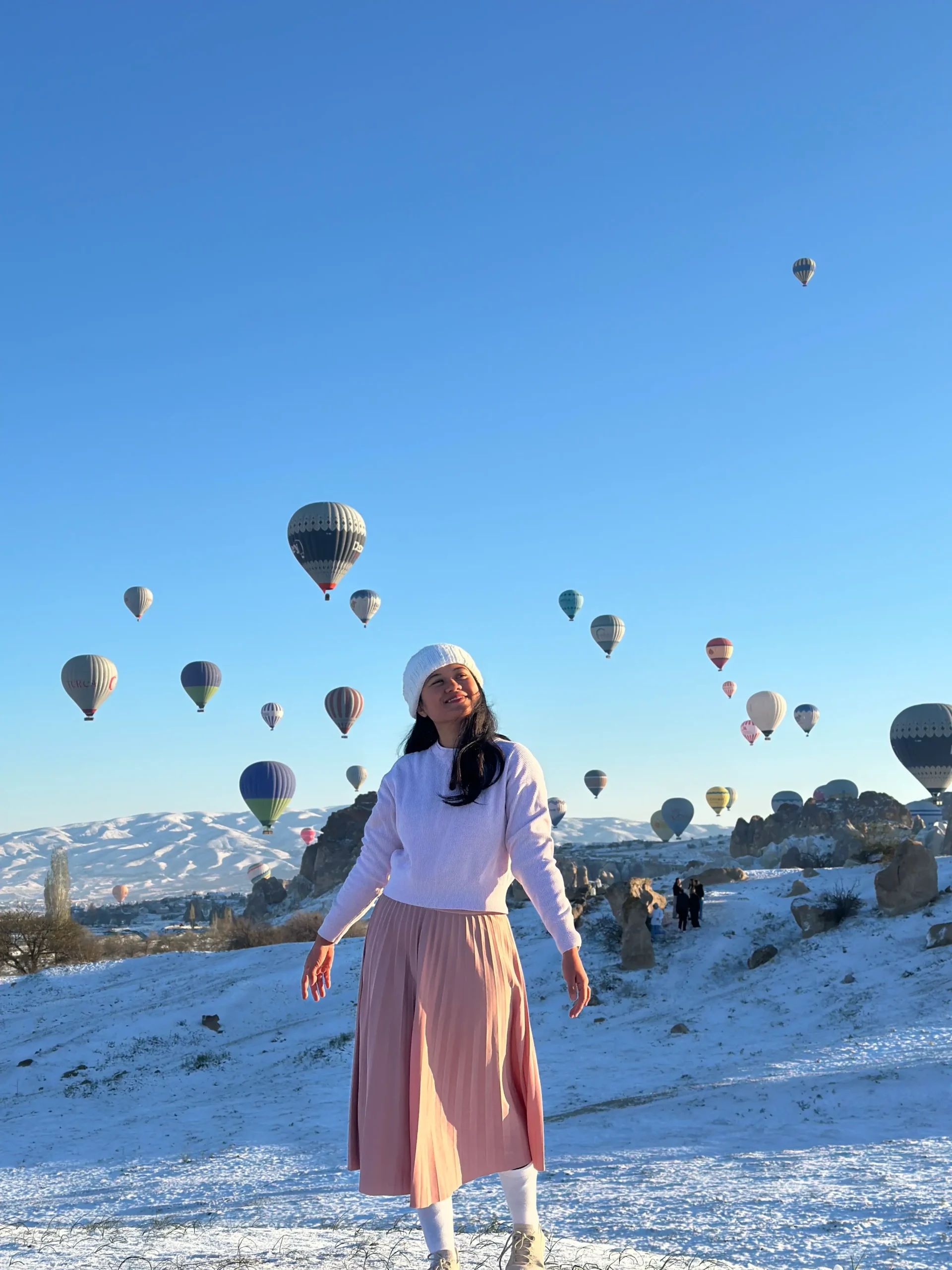 A woman in a white sweater and pink skirt stands in front of a blue sky full of hot air balloons during early winter, as part of her many work-exchange opportunities.