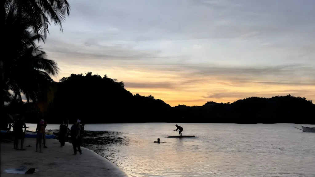 Silhouette of people at Doot Beach during sunset, with calm shallow water and trees in the background.