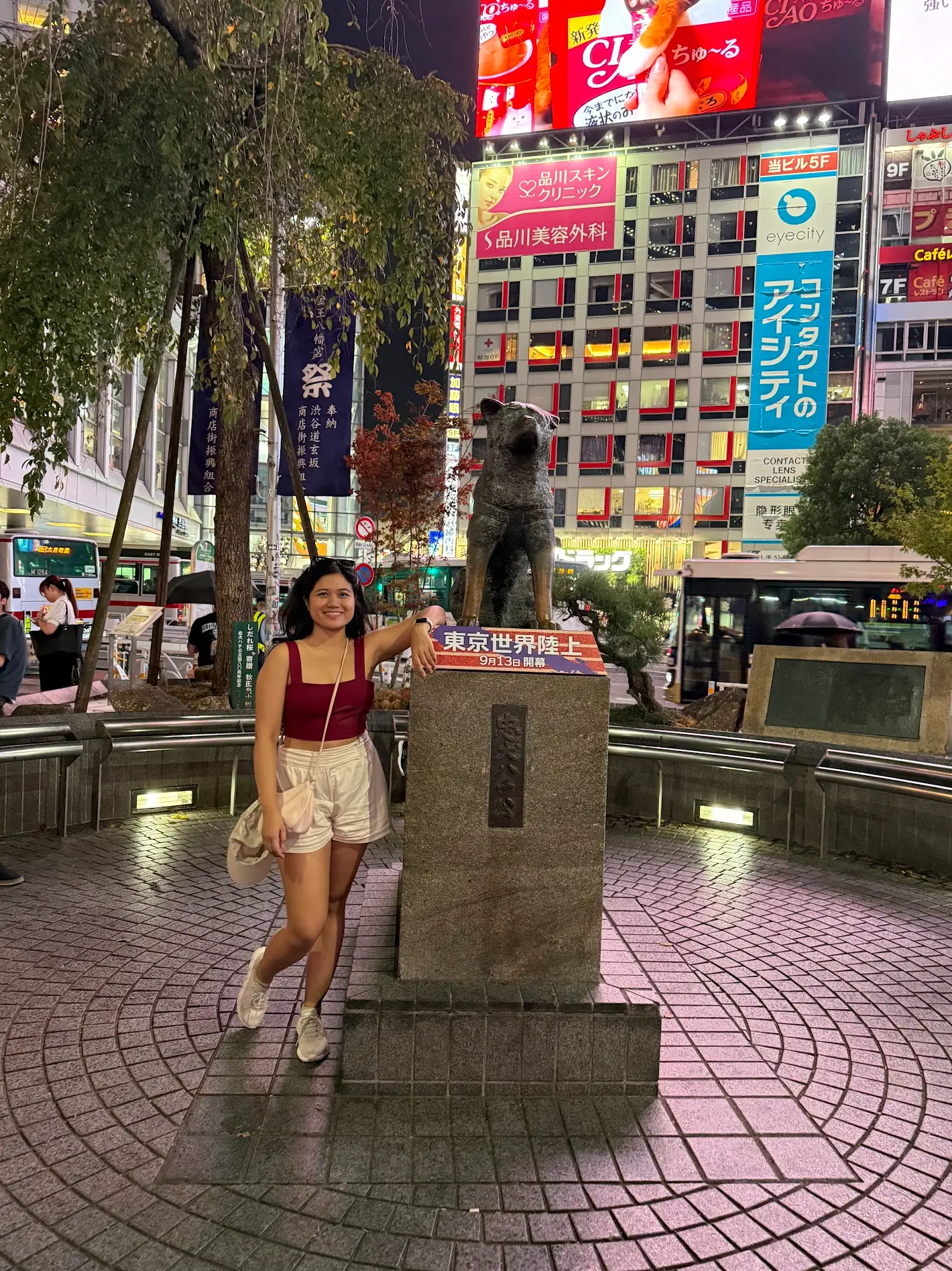 A woman standing beside the Hachiko statue in Shibuya at night with bright city lights and billboards in the background.