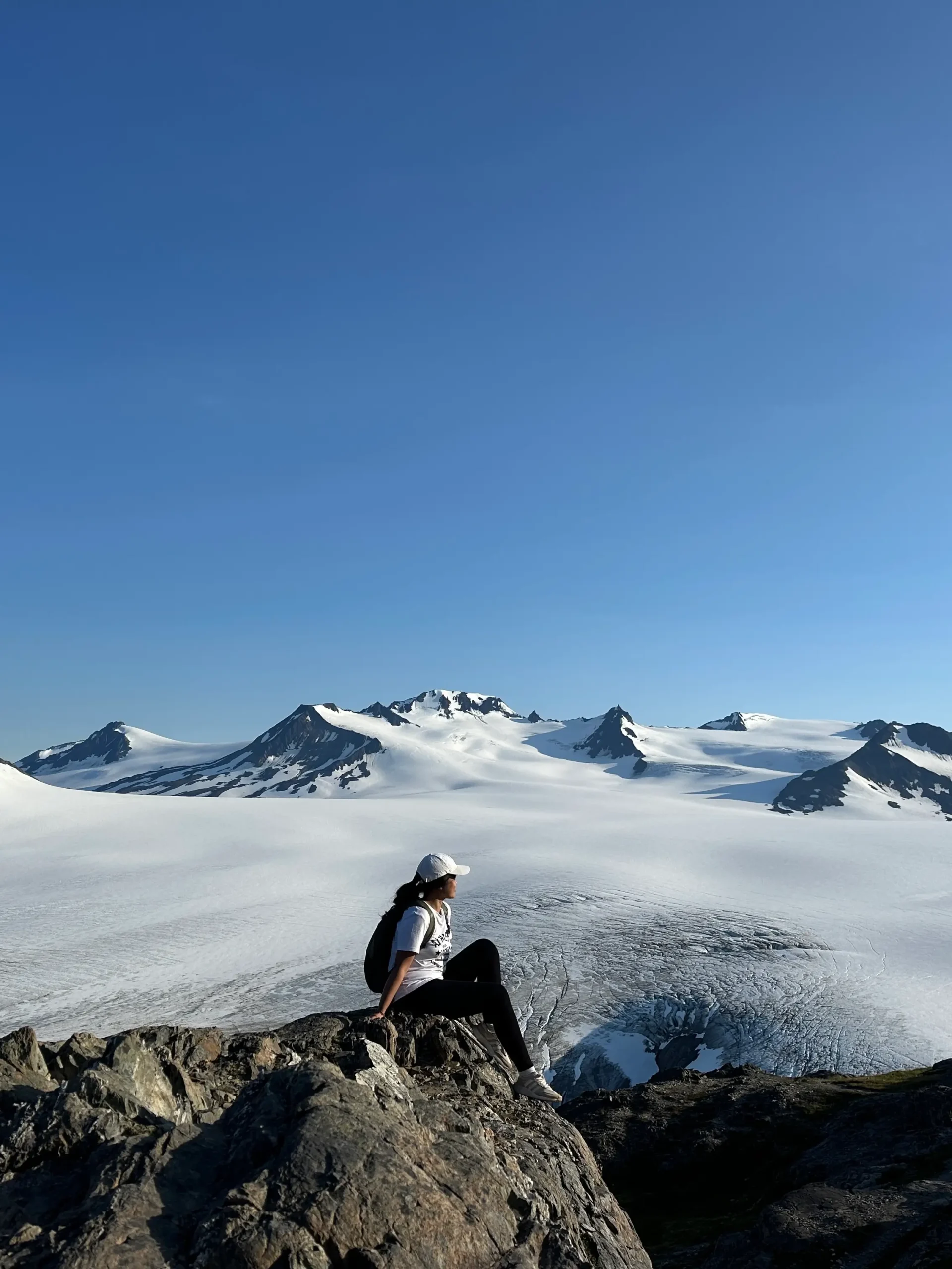 A woman sits at the top of a mountain in Alaska, surrounded by a clouds and other peaks.