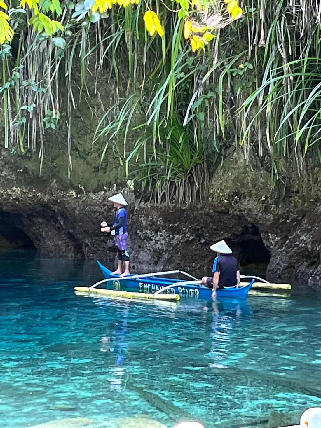 Two people wearing swimming gear and hats are on a blue boat, paddling deeper into the crystal blue waters and rock formations of Hinatuan Enchanted River, a hidden gem in Mindanao.
