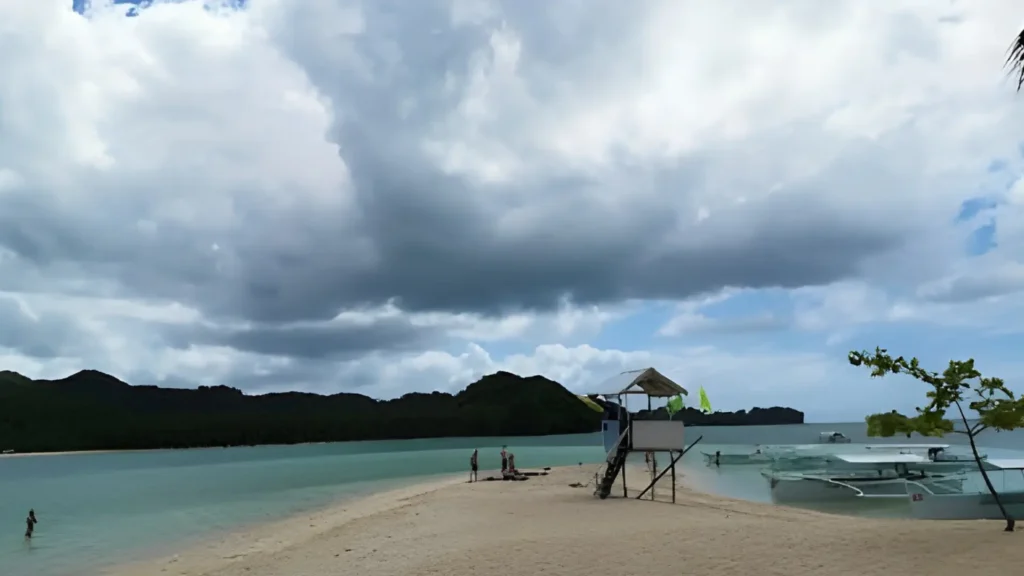 Kawhagan Island sandbar with shallow turquoise water, a small lifeguard hut, and boats anchored near the shore under a cloudy sky.