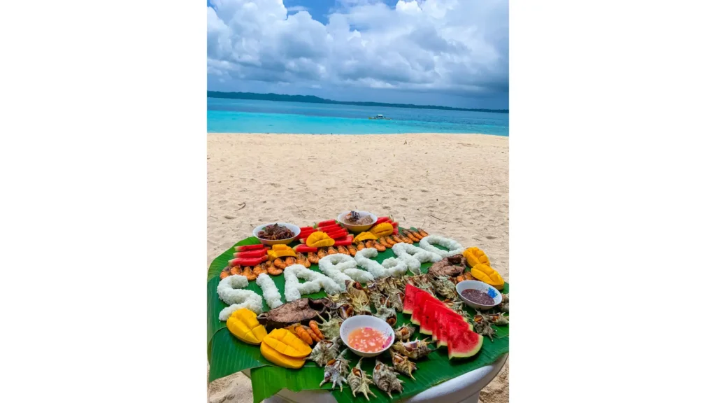 Seafood boodle meal arranged on banana leaves on the sand at Mam-on Island, with clear turquoise water and a wide beach in the background.