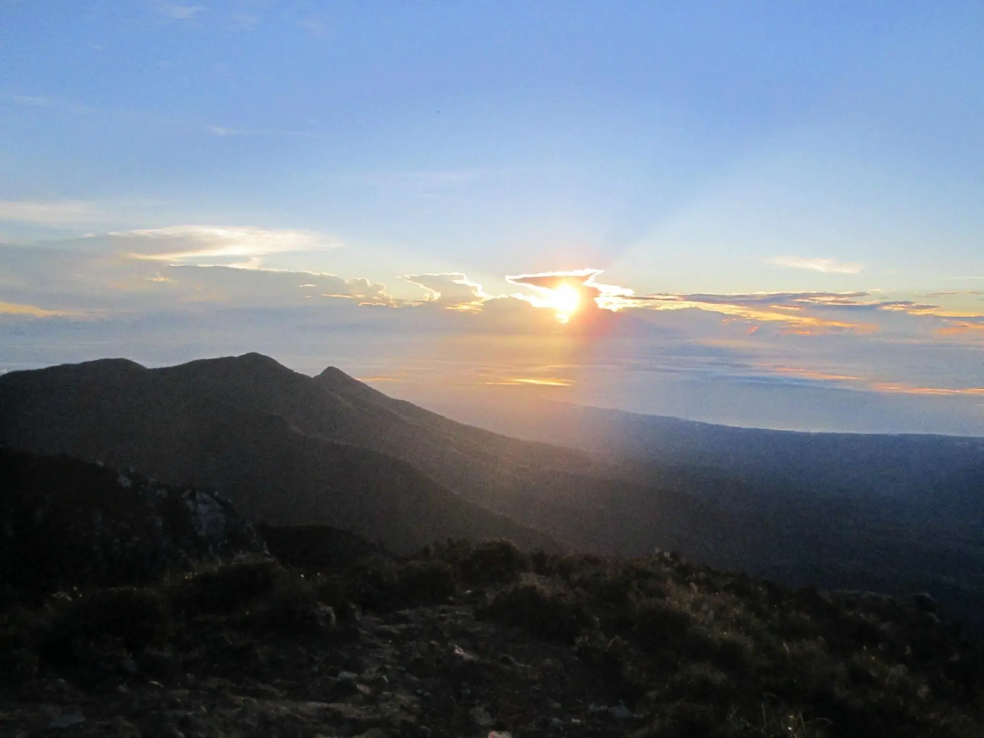The sun is partly hidden by the clouds, yet it casts an orange glow and long shadows over the landscape of Mt. Apo.