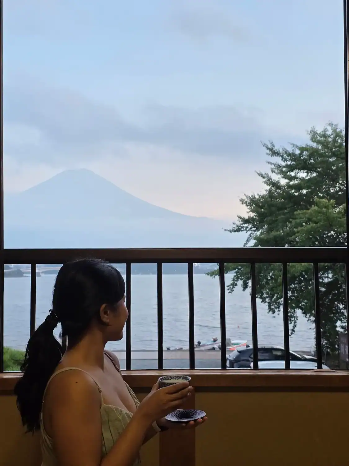 Traveler sitting inside a ryokan room, holding a cup of tea while looking out at Lake Kawaguchiko with Mount Fuji faintly visible behind the clouds.