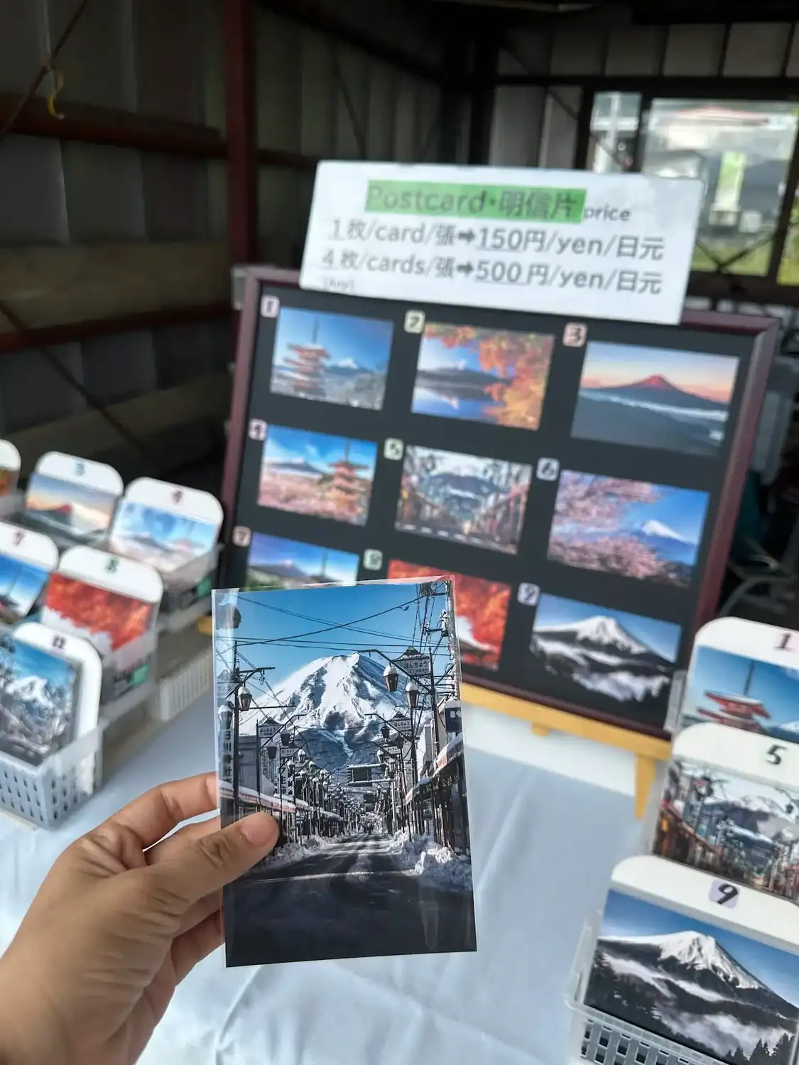 Hand holding a postcard of snowy Honcho Street with Mount Fuji in the background, displayed at a postcard stand in Fujiyoshida.