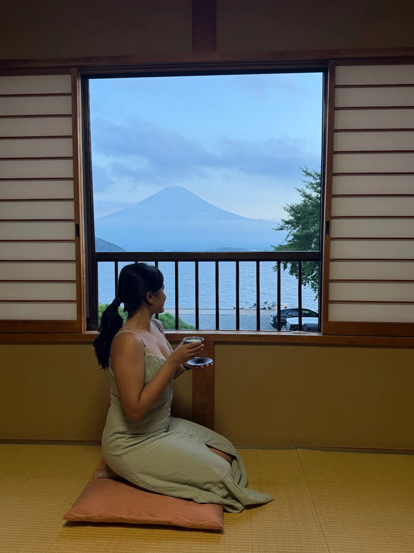 A woman sitting on a floor cushion in a tatami room while holding a cup and looking out at a lake and Mount Fuji through an open window.