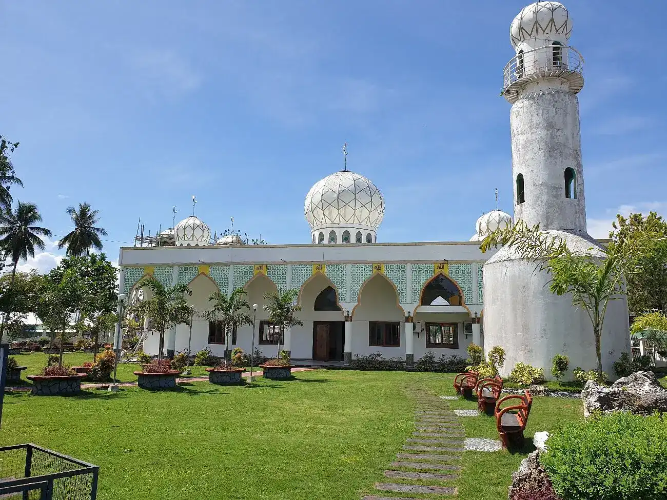 The facade of the Sheik Karim al Makhdum Mosque, featuring its white-painte tower and exterior, as well as the plant-lined road leading to the entrance.