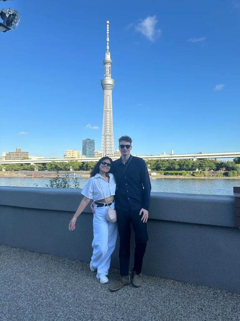 Two people standing together in front of the Tokyo Skytree with a clear blue sky and river behind them.