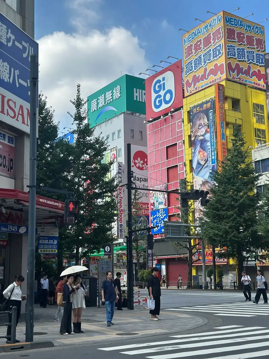 Busy street crossing in Akihabara, Tokyo, with colorful building signs, trees, and people waiting at the crosswalk on a sunny day.