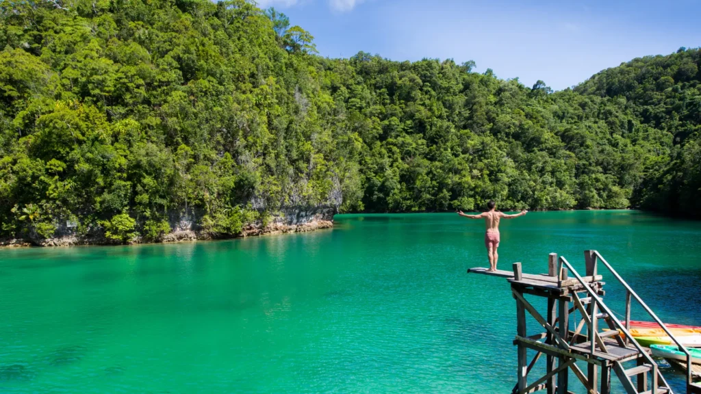 Person standing on a wooden diving platform over clear turquoise water at Sugba Lagoon, surrounded by green forested hills.