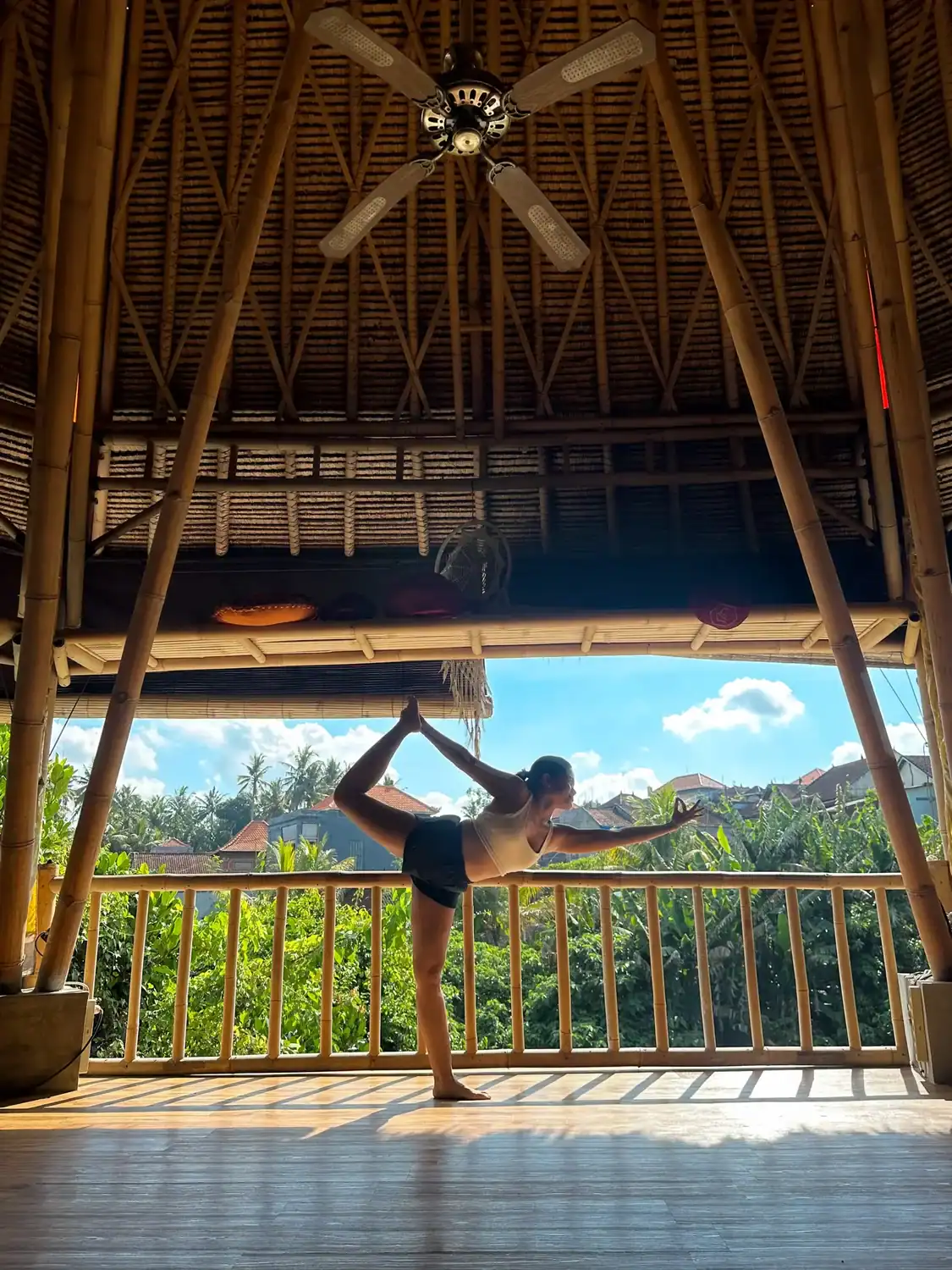 A woman is balancing on one leg while holding up the other in a backsplit. She is holding this position in a wooden hut with forests in the distance.