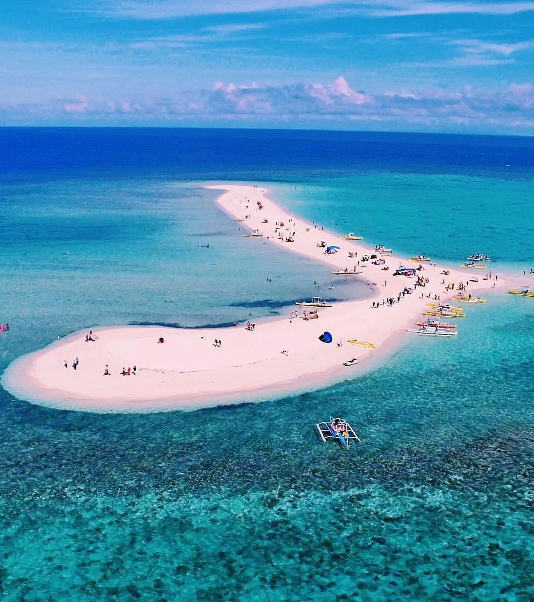 A strip of white sand emerges from a turquoise ocean, with people dotting the area. 