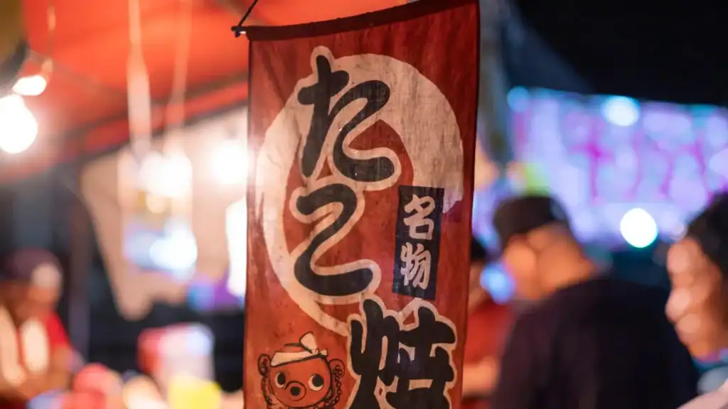 A takoyaki stall banner hanging at a Japanese summer festival with blurred vendors and bright lights in the background.