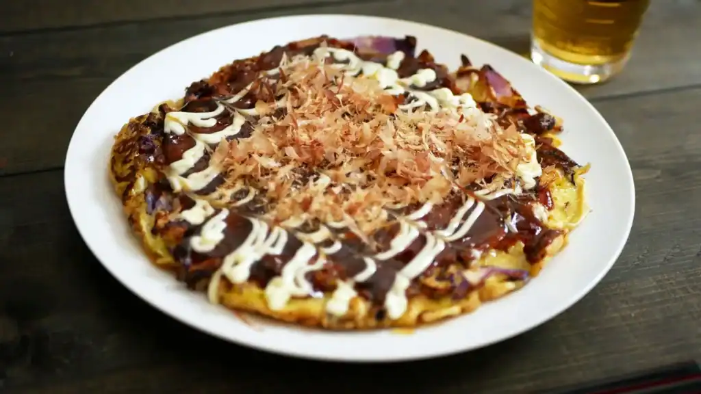 Osaka-style okonomiyaki topped with bonito flakes, mayonnaise, and savory okonomiyaki sauce on a white plate.