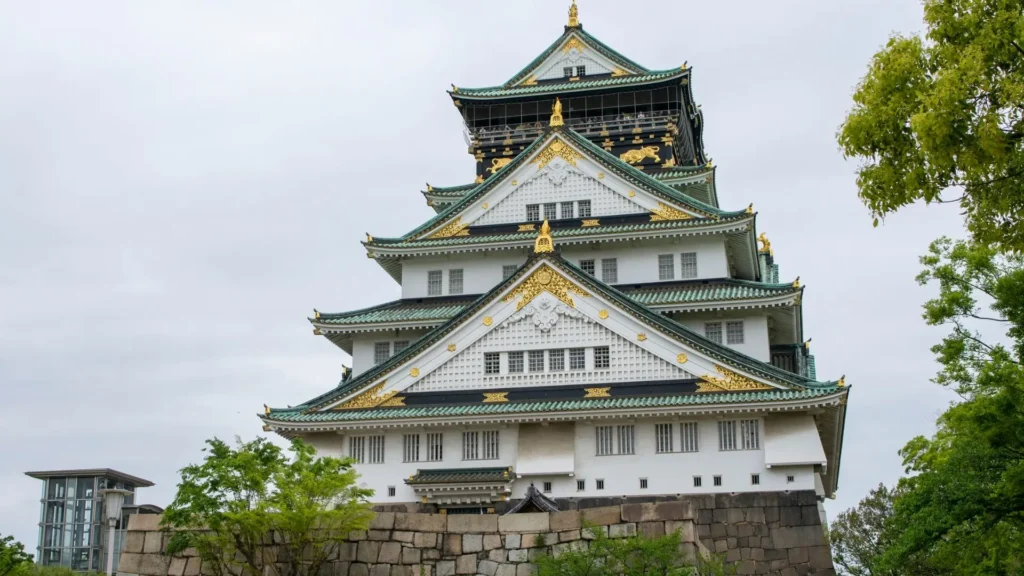 Things to Do in Osaka at Osaka Castle featuring white walls green tiled roofs gold details stone walls and surrounding trees.