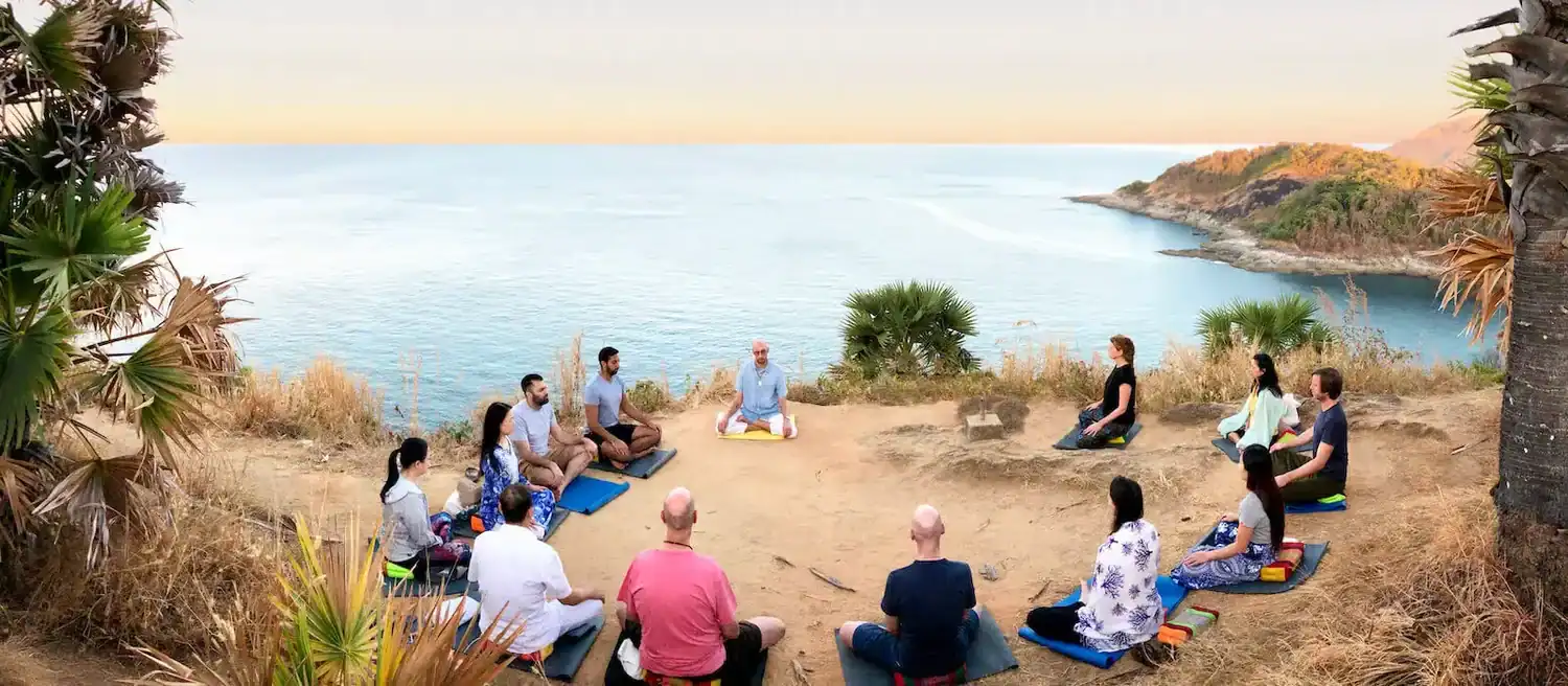 A group of people sit in a circle of a cliff overlooking an ocean, as part of a free meditation session held by the Phuket Meditation Center, one of the best wellness retreats in Phuket.