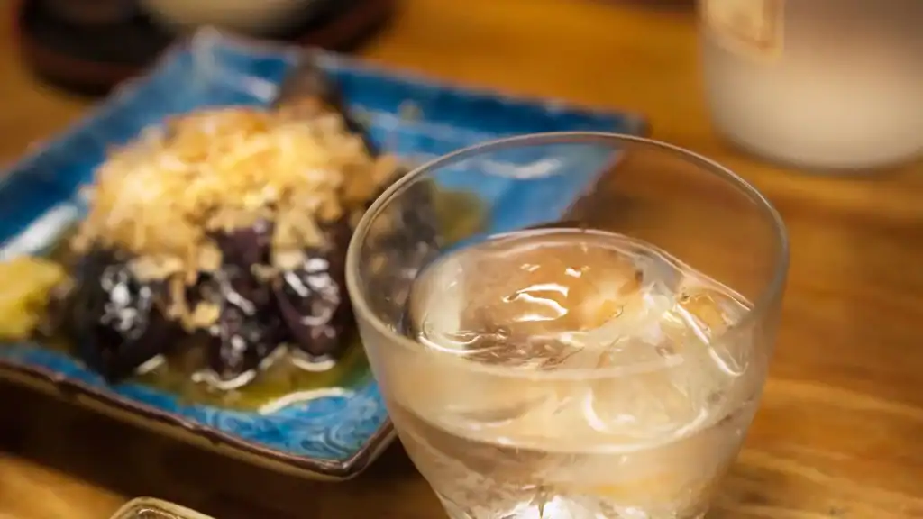 Glass of Japanese sake served on ice beside a plate of izakaya appetizers, showcasing traditional Japanese drinks and food to try in Japan.