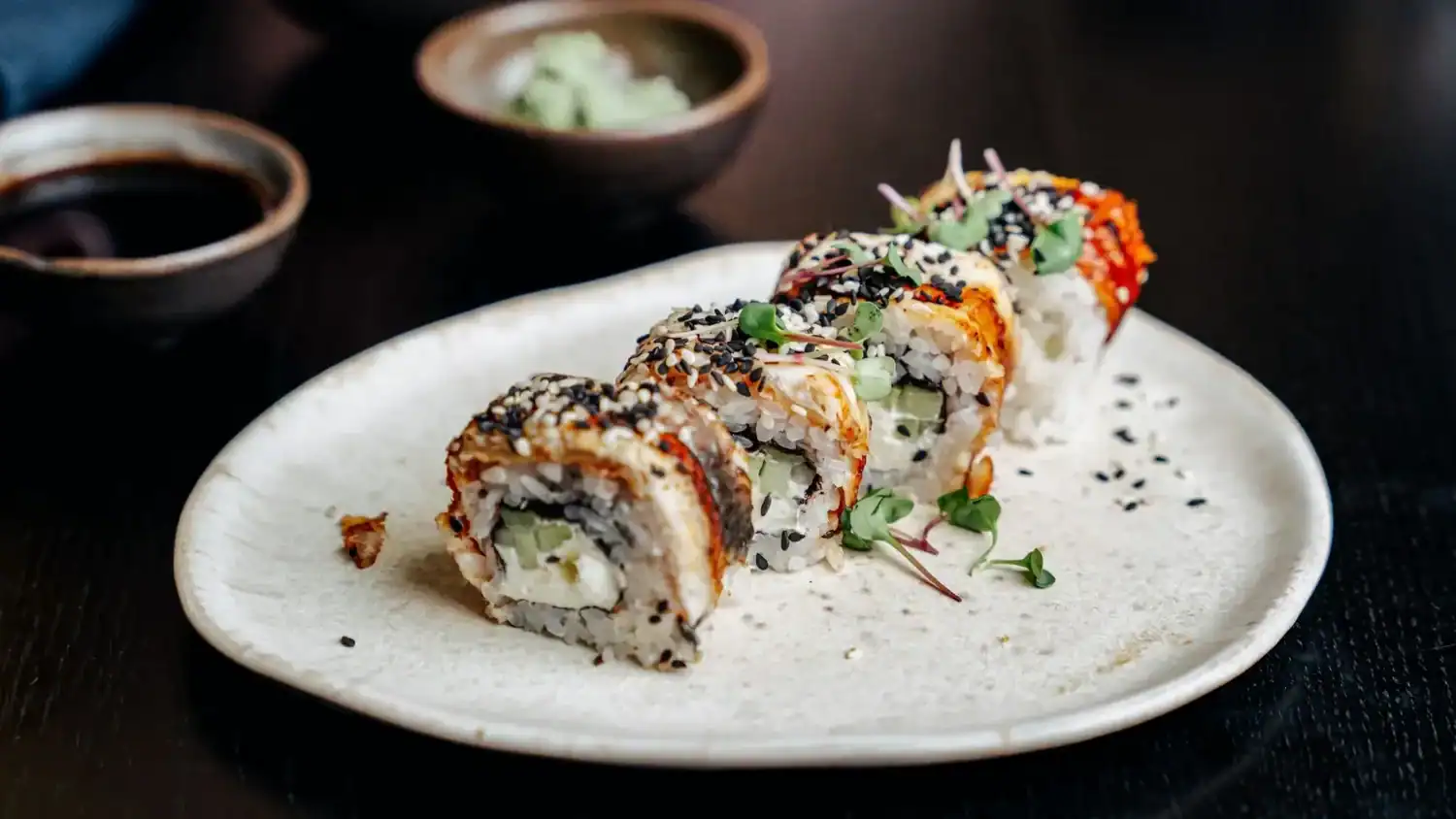 Japanese sushi roll topped with sesame seeds and microgreens, served with soy sauce and wasabi.